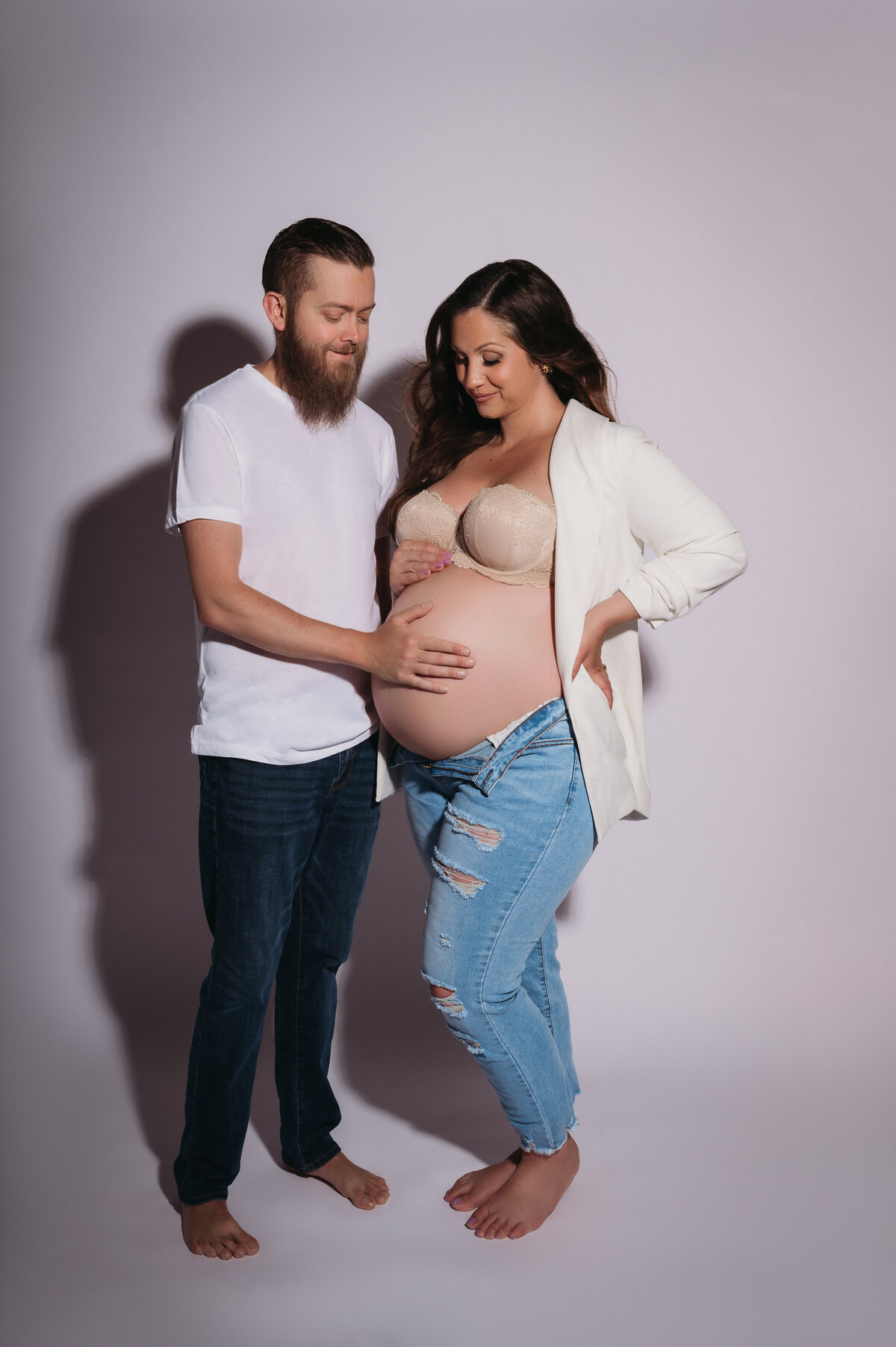 parents to be looking down and smiling at mom to be's baby bump with mom wearing a white blazer and jeans and dad wearing a white t shirt and jeans in front of a white backdrop