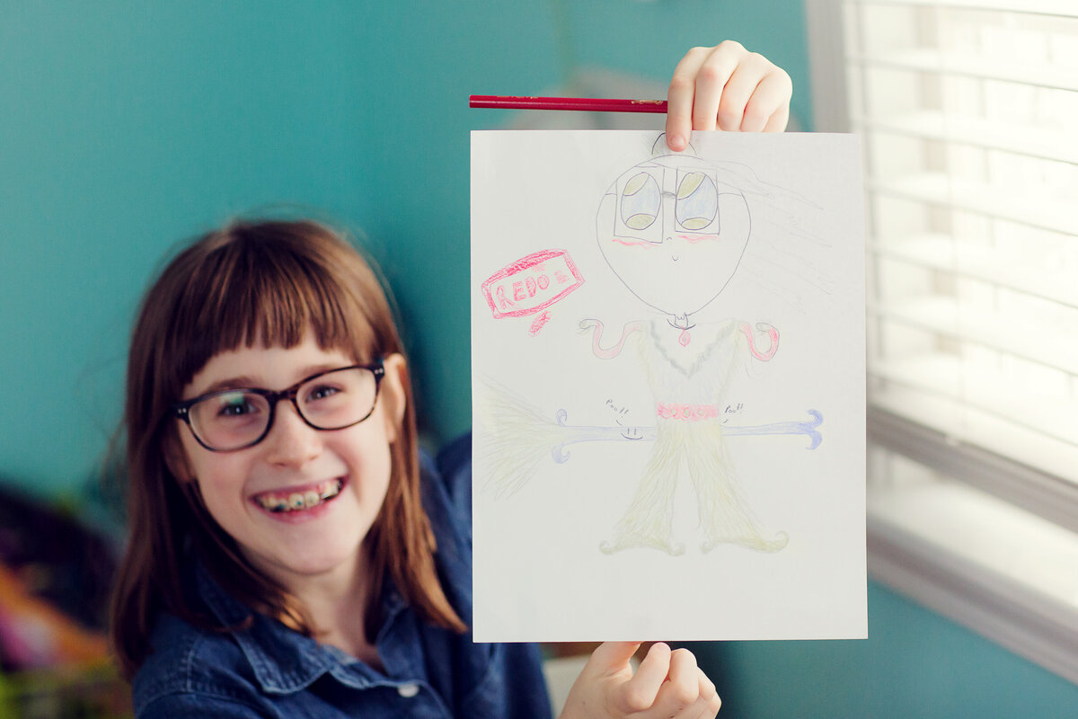 A girl happily shows off her drawing. She has on glasses and the walls behind her are blue.