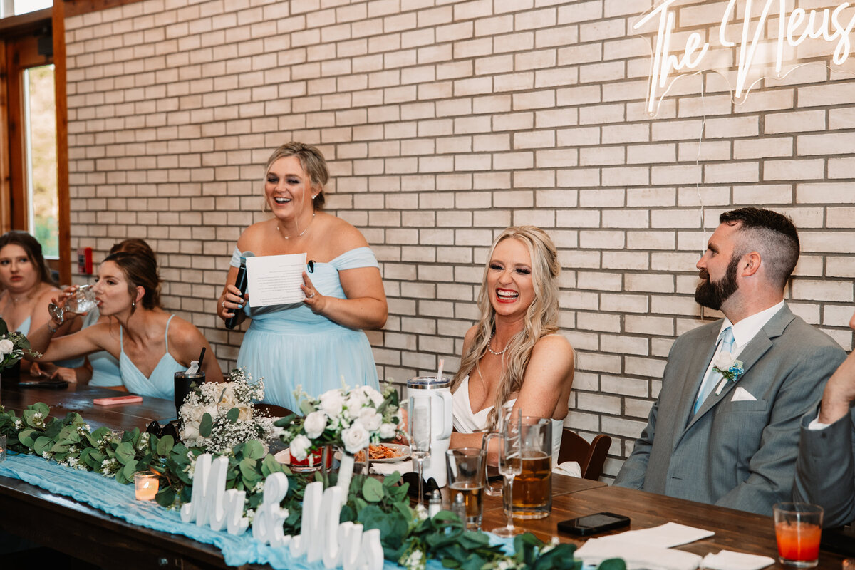 A bridesmaid in a blue dress gives a toast while bride laughs at long head table against brick wall