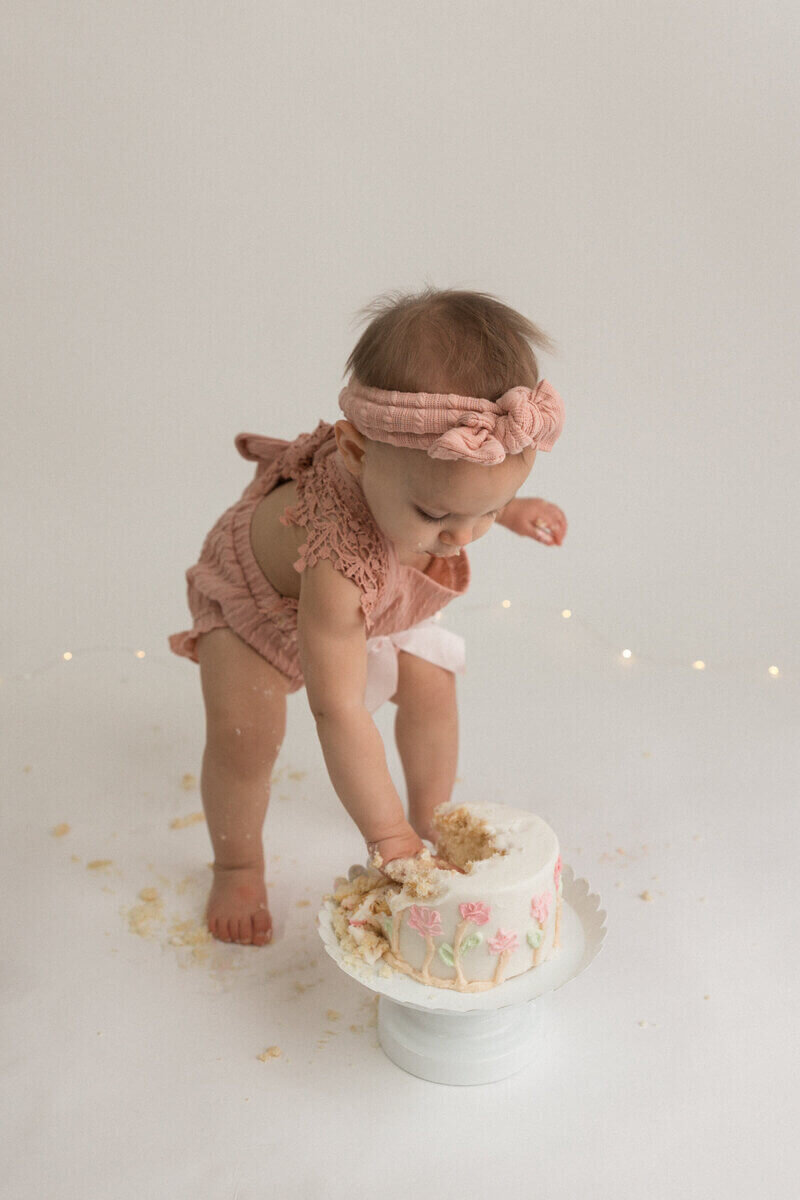 A baby girl in a pink outfit leans forward, digging her hand into a half-eaten cake with pink floral decorations. Cake crumbs are scattered on the white floor around her.