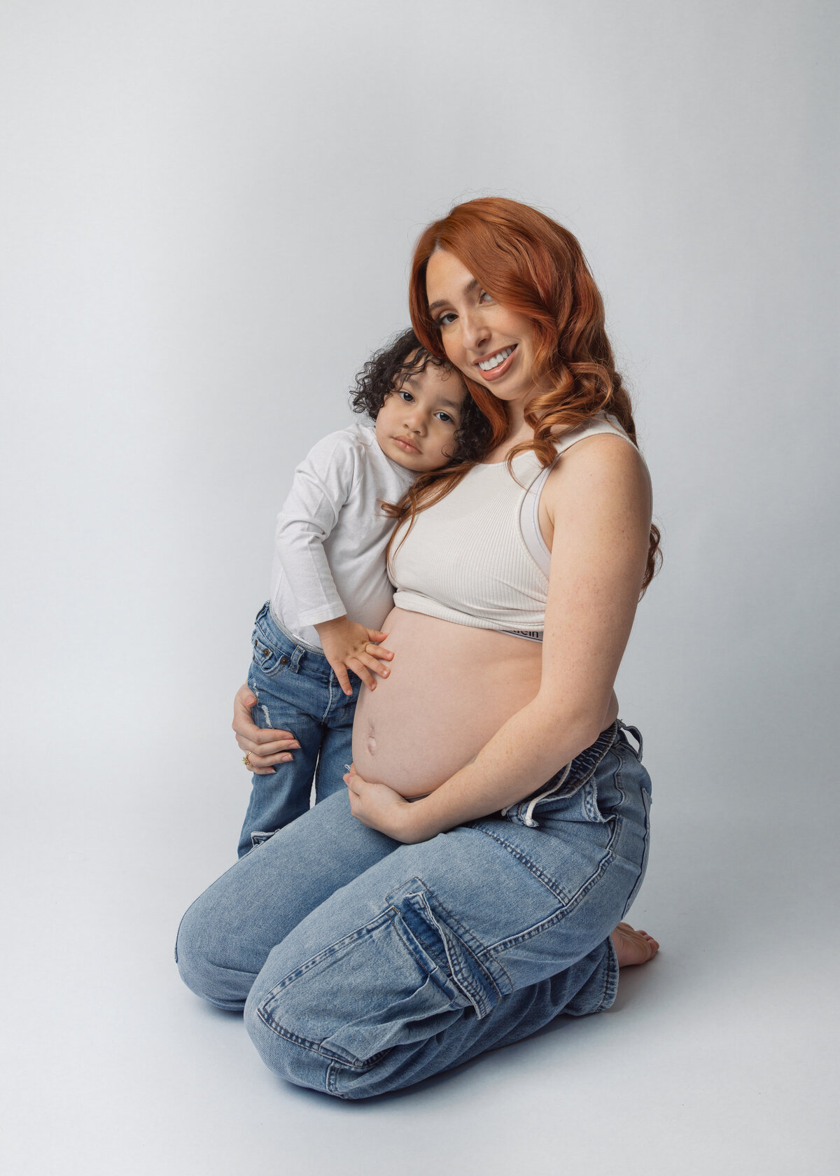 Pregnant women in jeans and a white crop top hugs her young toddler during a maternity session in North Jersey.