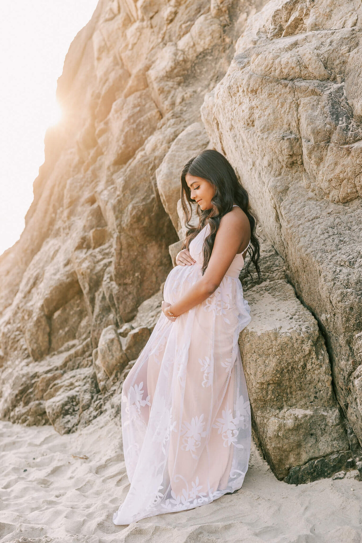Woman with long black hair leaning on rocks and smiling down at her pregnant belly. She is wearing a long white dress with no sleeves on it and the sun is peeking over the edge of the rocks on the beack.