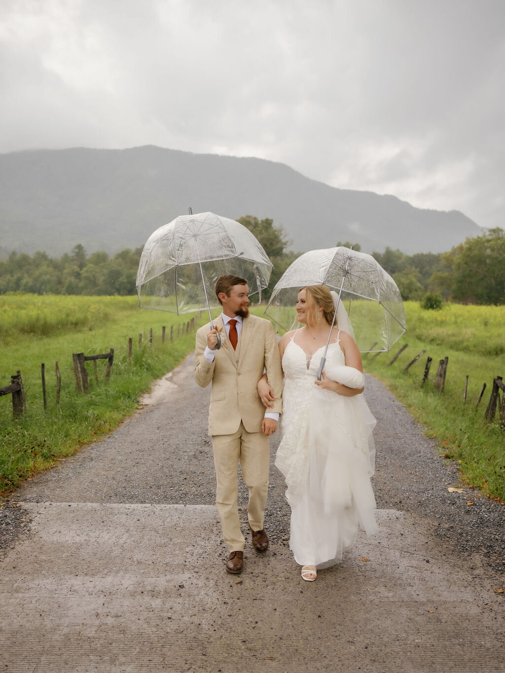 bride and groom hold hands and walk down a cement path in Cades Cove for their Smoky Mountain elopement while holding clear umbrellas to stay dry from the rain