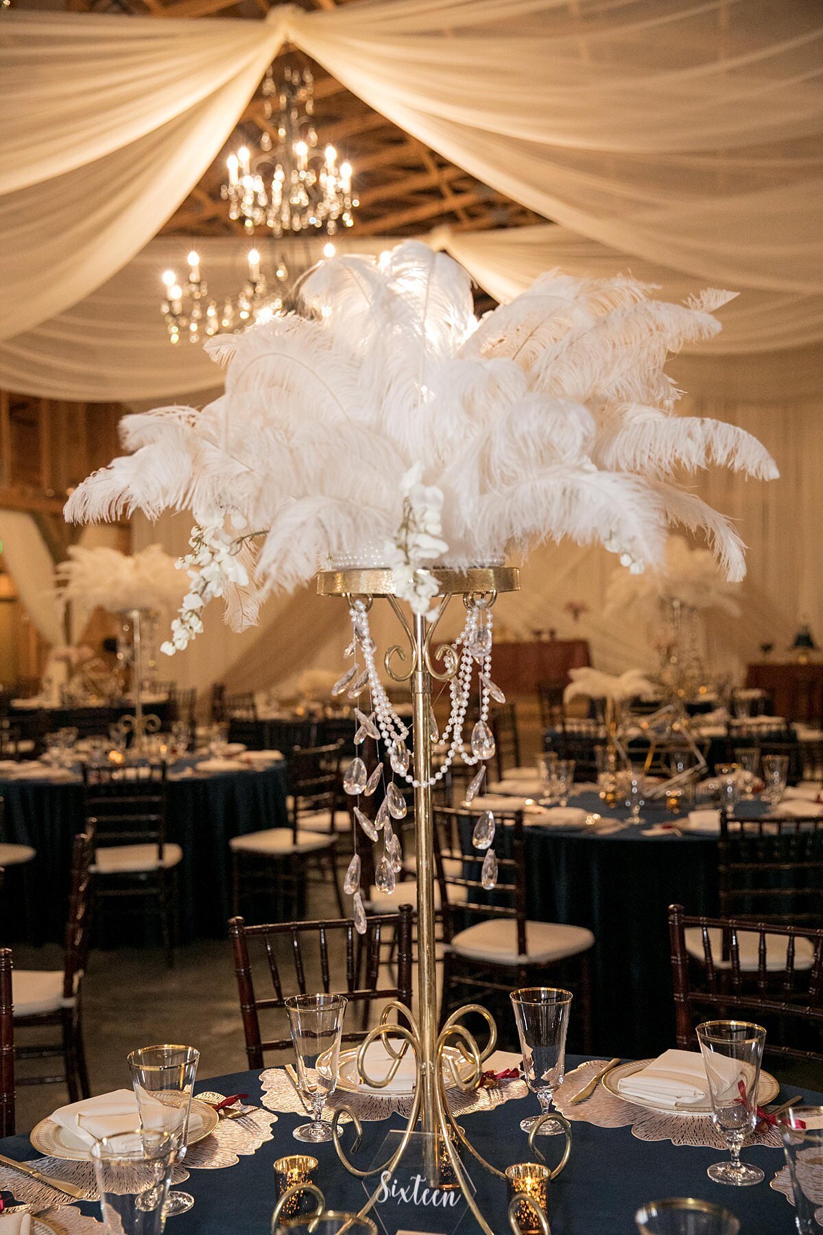 Tall ostrich feather centerpiece accented with hanging crystals on a tall gold stand. The table is set with a navy blue velvet table cloth, footed crystal water goblets with gold rims, ginko leaf chargers and ivory china plates with gold accents. The matte gold flatware has a small thank you note tied to each fork and there are gold votive candles on each table. Brown chiavari chairs with ivory cushions are placed around each table and the ceiling is covered in sheer ivory drapery accenting the gold and crystal candelabra chandeliers of the cedar wood barn at Saddle Woods Farm.