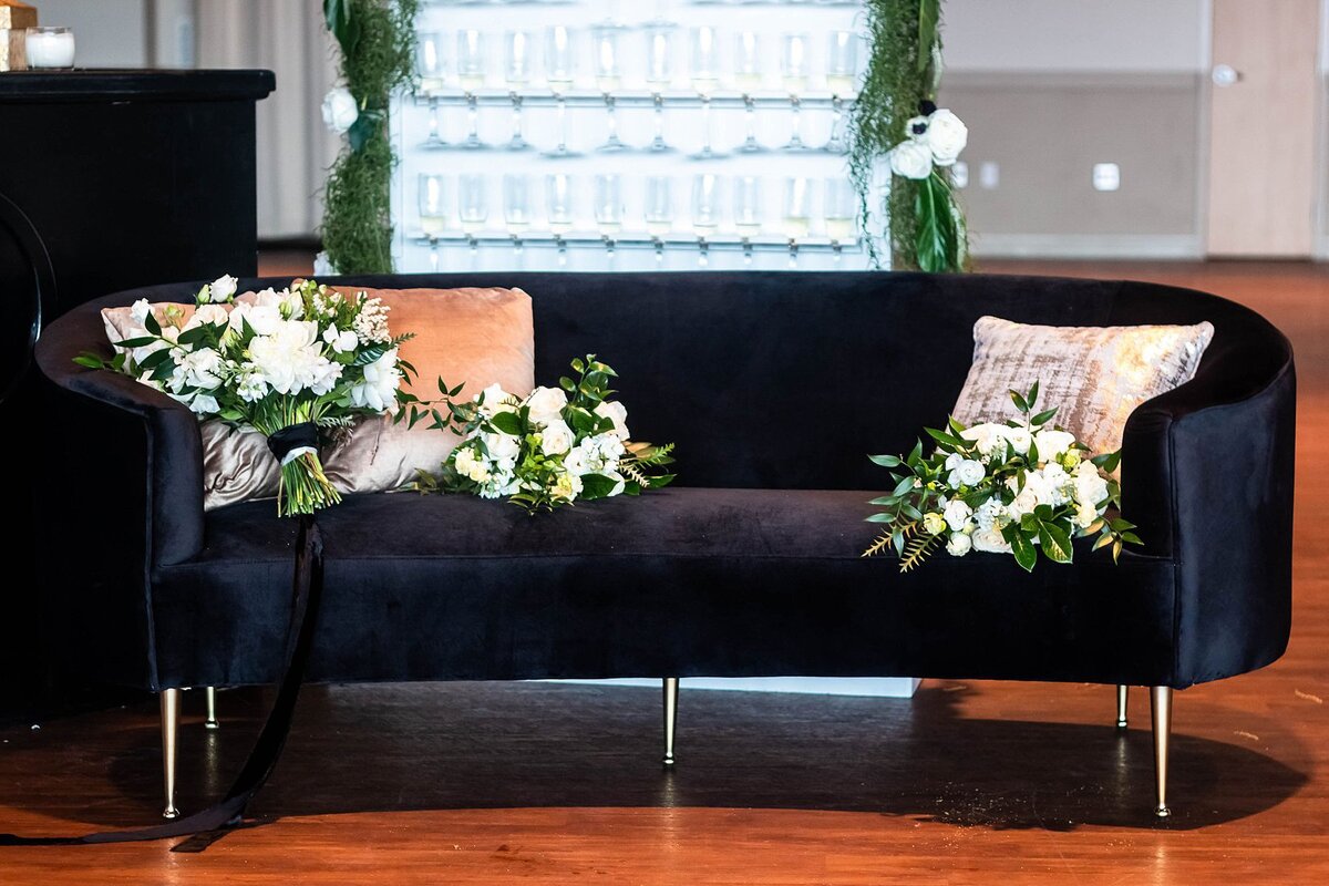 A mid century modern sleek black sofa on thin silver legs sits in front of a champagne wall at a Noah Liff Opera Center wedding. The sofa has gold and rose gold sequined throw pillows and three large white floral bouquets with greenery.