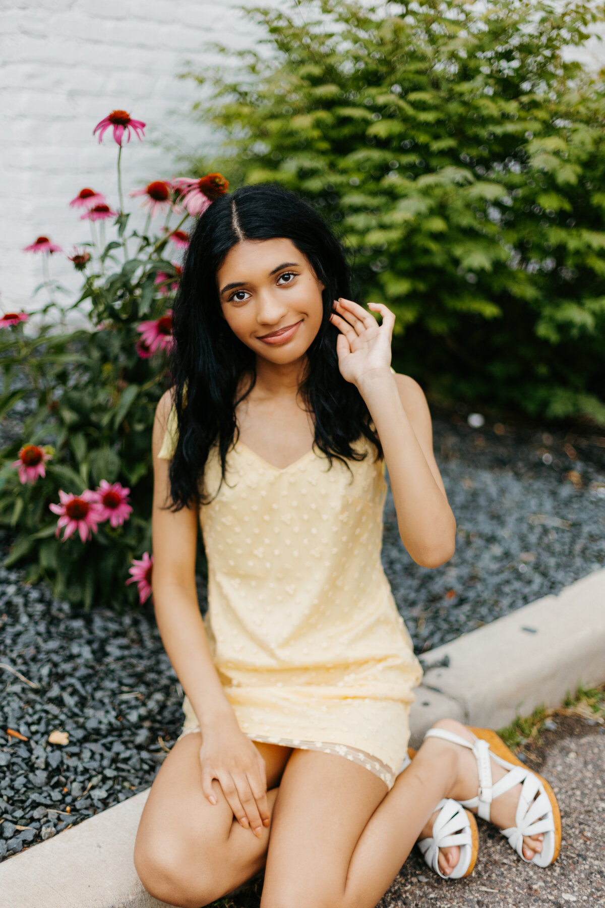 Senior girl in a yellow dress sitting on the ground in front of gravel and pink flower bushes