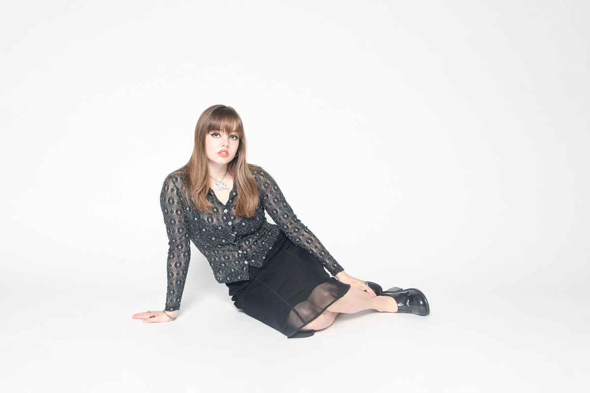 A person with long hair is sitting on the floor against a white background. They are wearing a patterned sheer top, a black skirt, and black shoes. The pose is relaxed, with one hand on the floor and legs crossed.