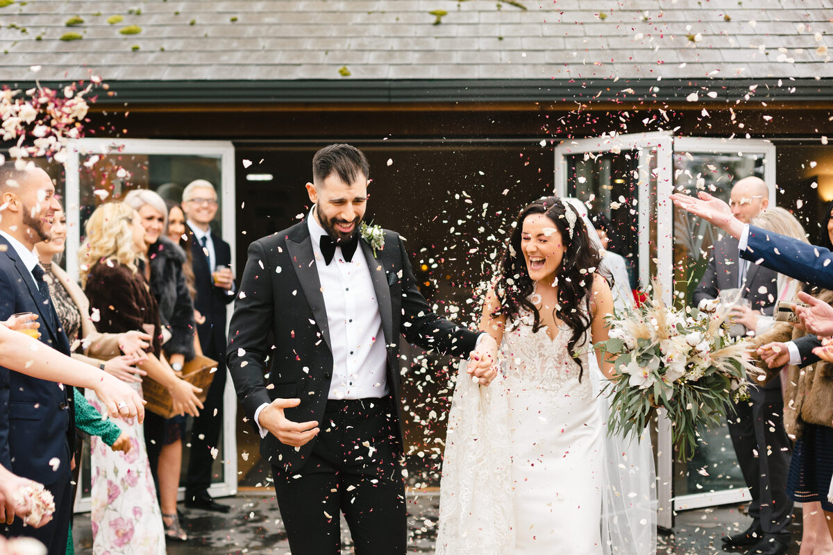 A newlywed couple, smiling and holding hands, walk outside as guests shower them with confetti at their Wasing Park Wedding. The bride wears a white dress and holds a bouquet, while the groom looks sharp in a black suit. People celebrate around them.
