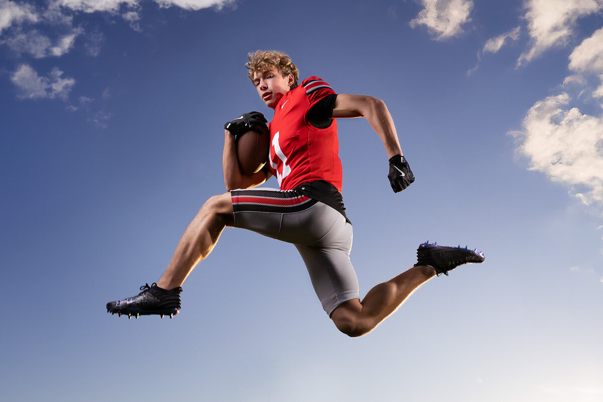 Senior football player in red uniform mid-leap with football, showcasing athleticism in action, captured for Dallas senior photos. The dynamic shot highlights his strength and determination.