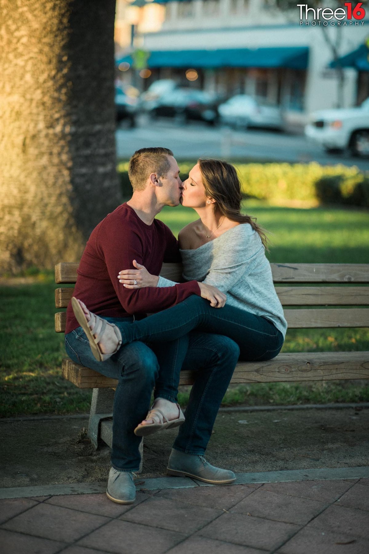 Engaged couple sit on top of each other on a bench as they kiss