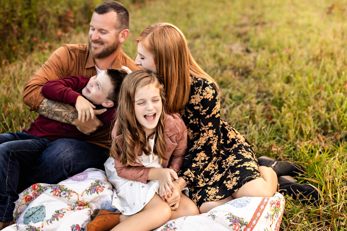 A family enjoys a sunny afternoon on a patterned blanket in a grassy field. Captured in vivid outdoor portraits by a Bloomington family photographer, the father, wearing a brown shirt, holds their giggling boy while the mother in her floral dress smiles warmly at their daughter in white and pink.