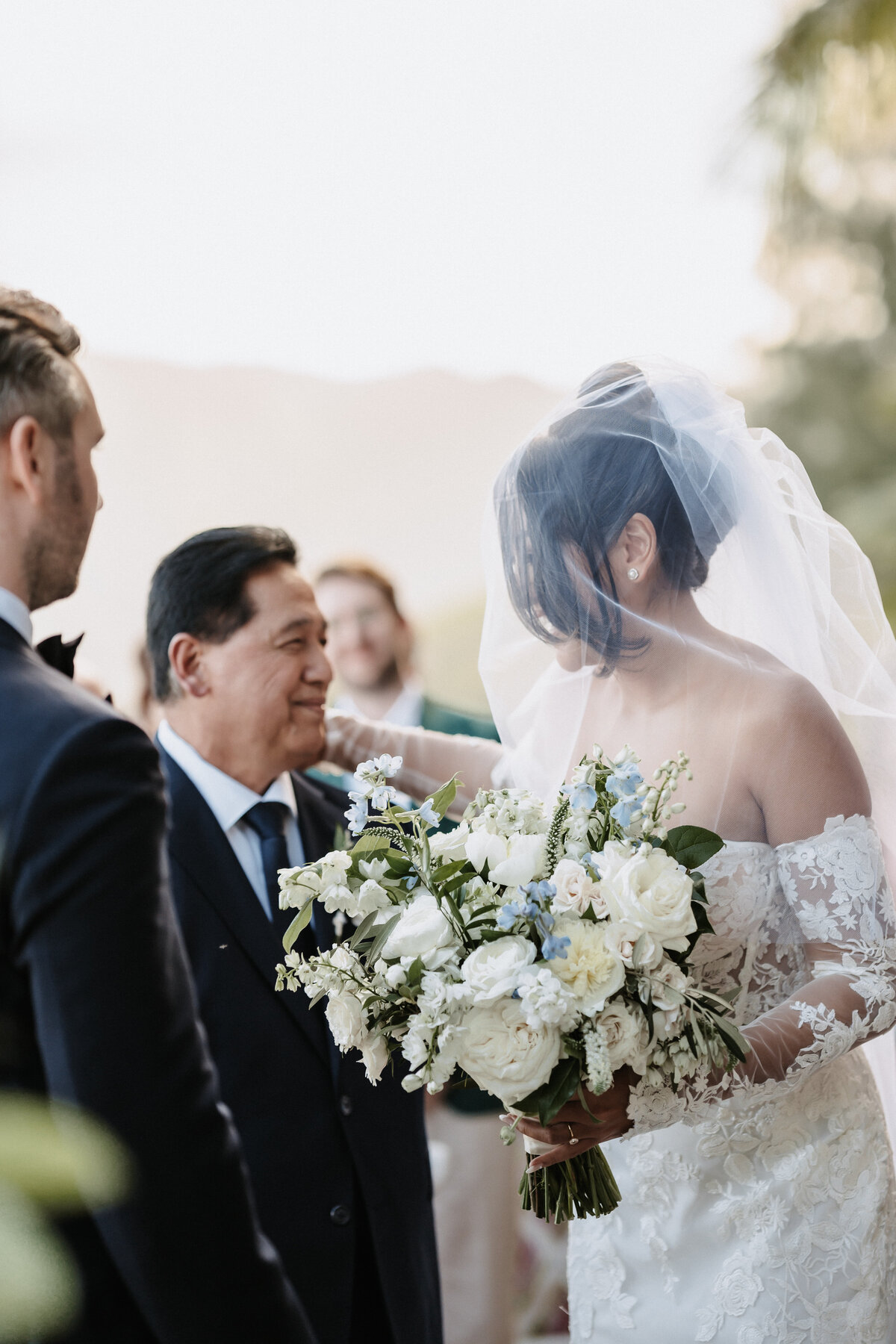 Bride looks lovingly at her father as he hands her off to groom while she holds flowers in lace wedding dress and veil over her head