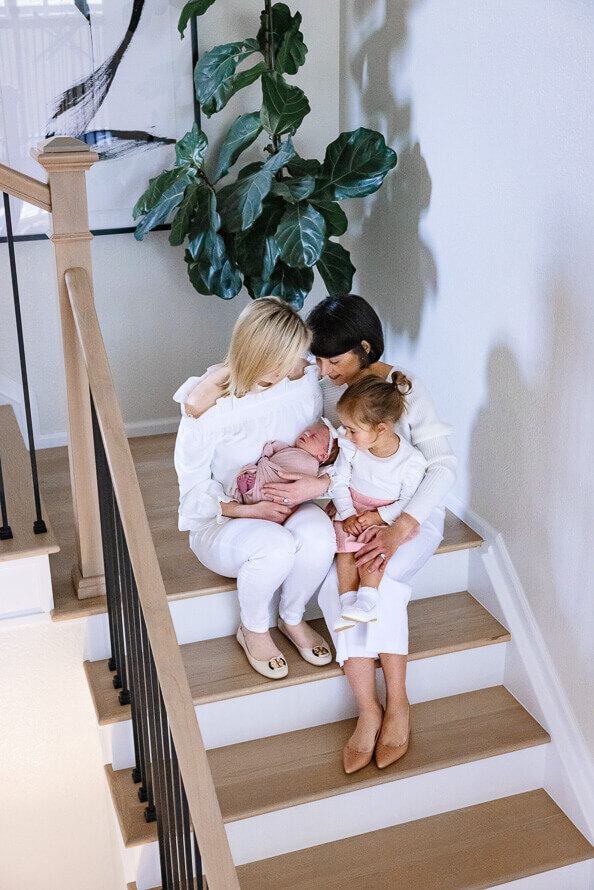 Two moms and toddler girl snuggle on stairs and look at their baby.