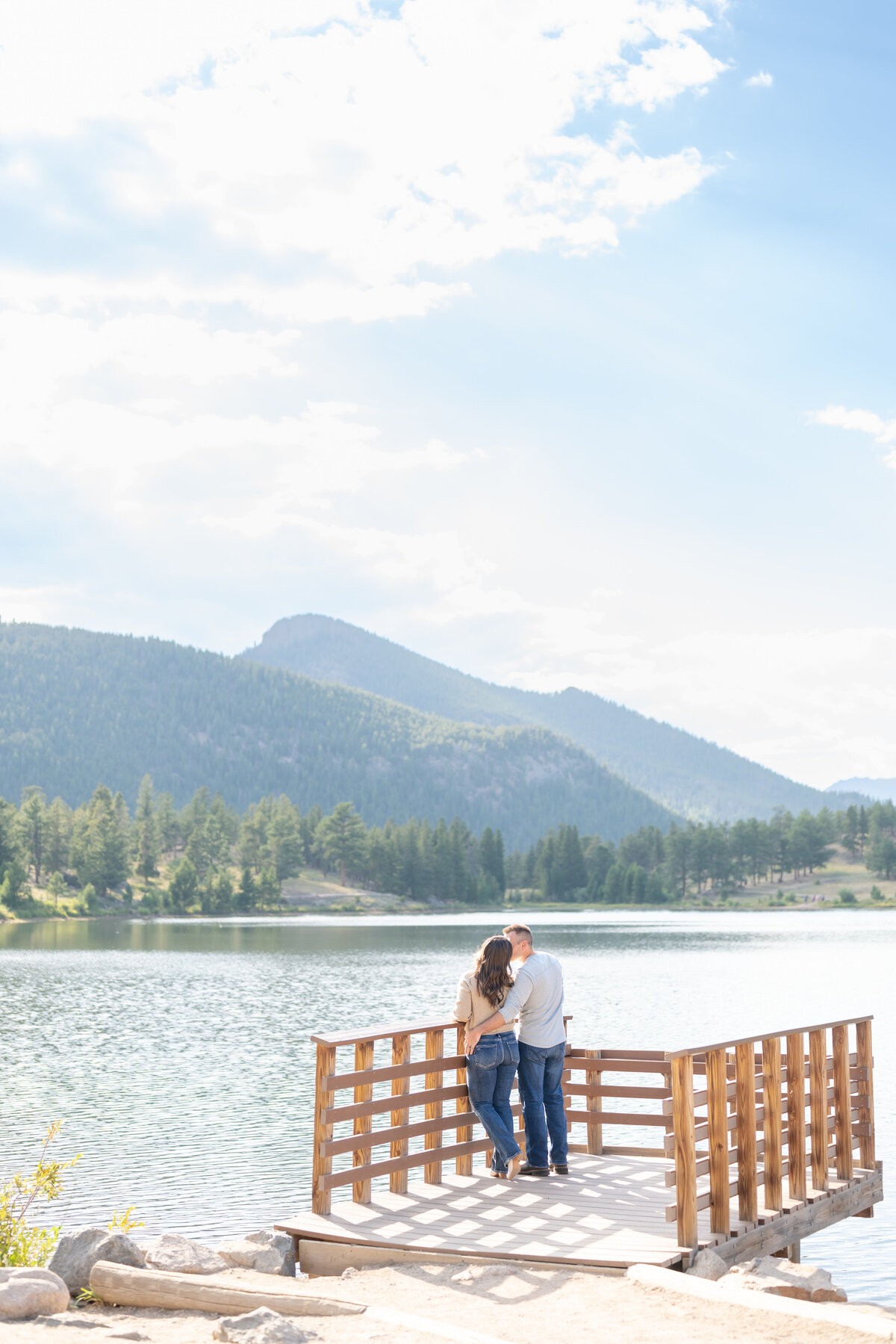 Couple leaning against wooden fence while kissing by the blue lake and mountain views in rocky mountain national park in Colorado