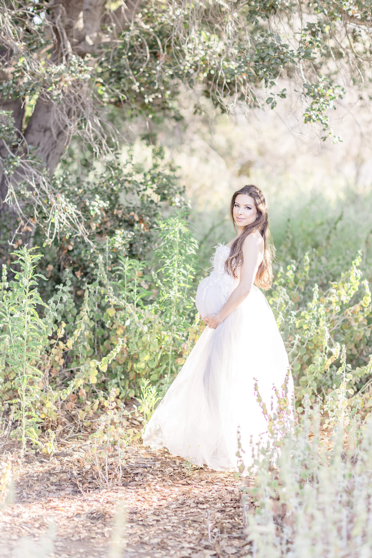 A pregnant woman stands in a sunlit forest clearing, wearing a flowing white dress. She cradles her belly and smiles softly, surrounded by lush greenery and dappled sunlight filtering through the trees.