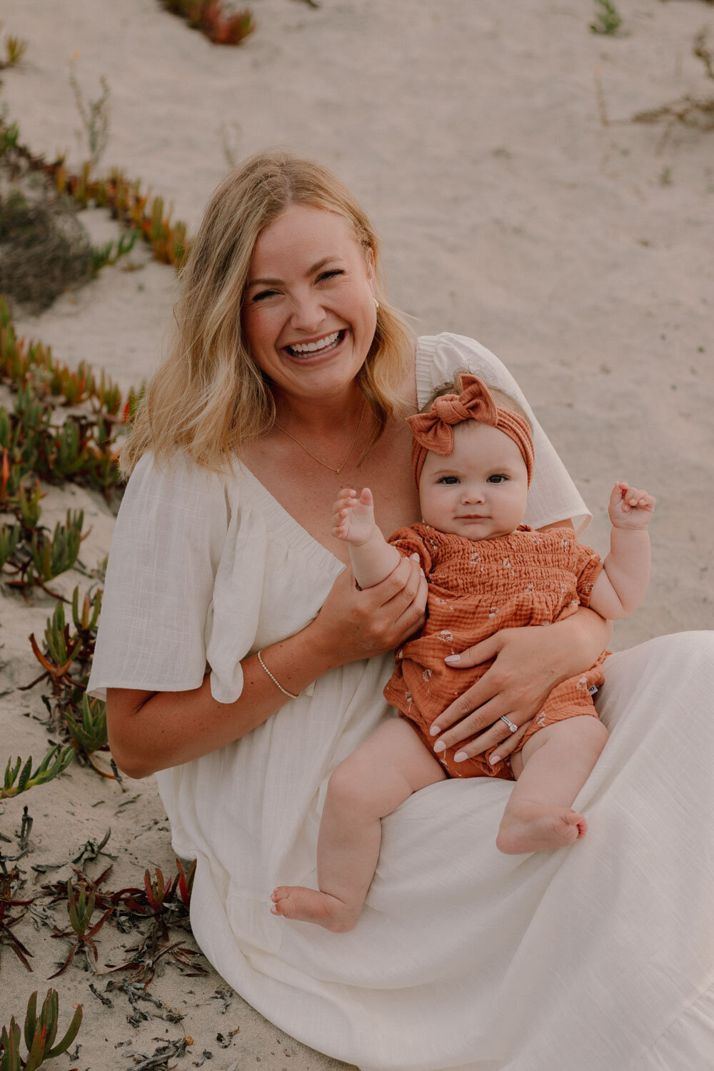 Mother smiling joyfully with her baby sitting on her lap on the beach, surrounded by greenery
