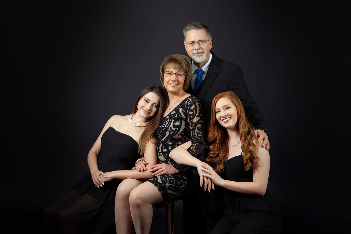 A family portrait captured by a Jacksonville family photographer showcases two adults standing behind two seated young women. They are all dressed in coordinated black attire against a dark backdrop, exuding a content and close-knit vibe.