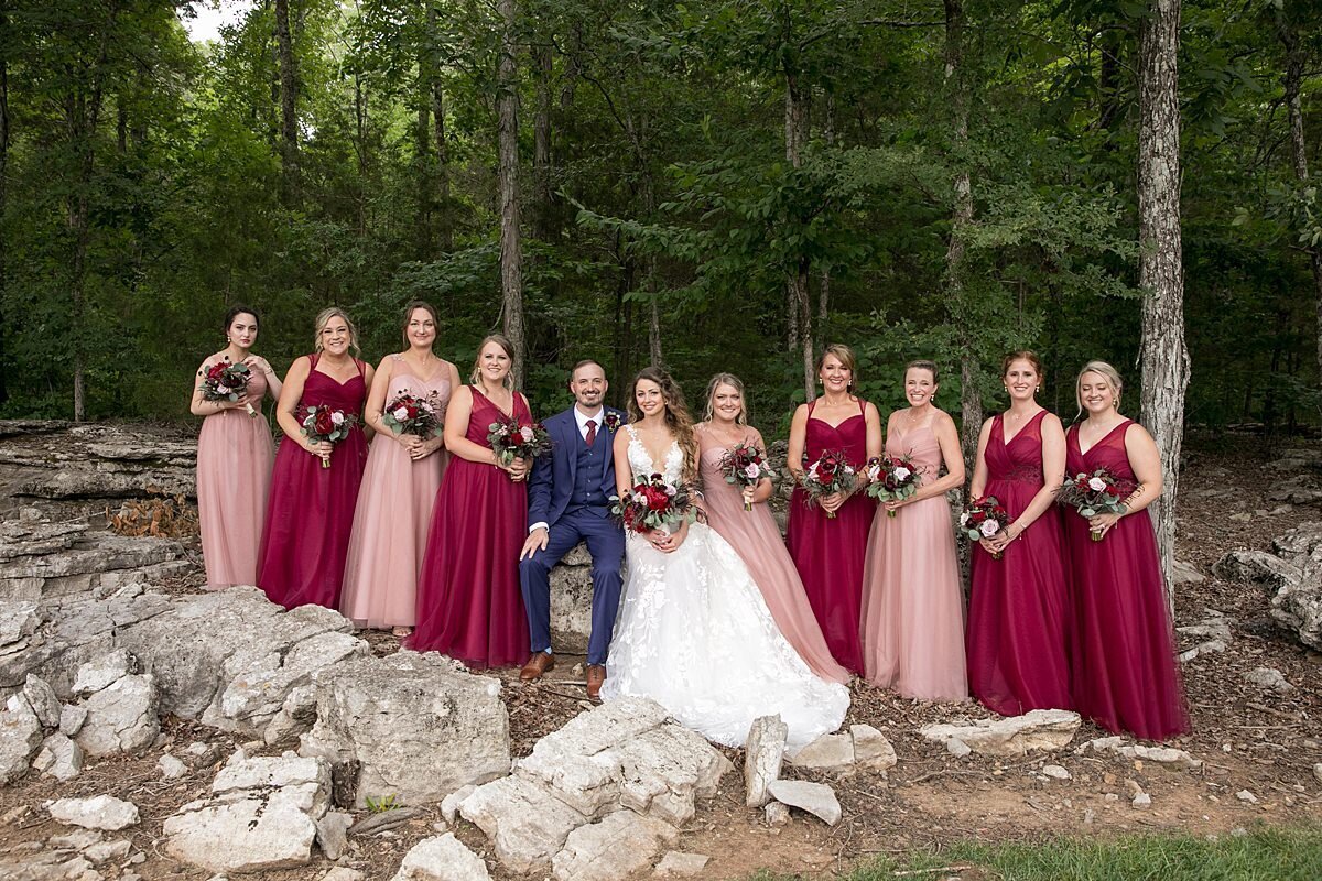 The bride, wearing a long lace wedding dress with a plunging neckline stands next to the groom wearing a navy blue suit with a burgundy tie as they pose on a limestone rock outcropping with the bridesmaids who are dressed in blush bridesmaid dresses and burgundy bridesmaid dresses while holding blush and burgundy bouquets of roses, ranunculus and peony wrapped in ivory satin ribbon at Saddle Woods Farm.