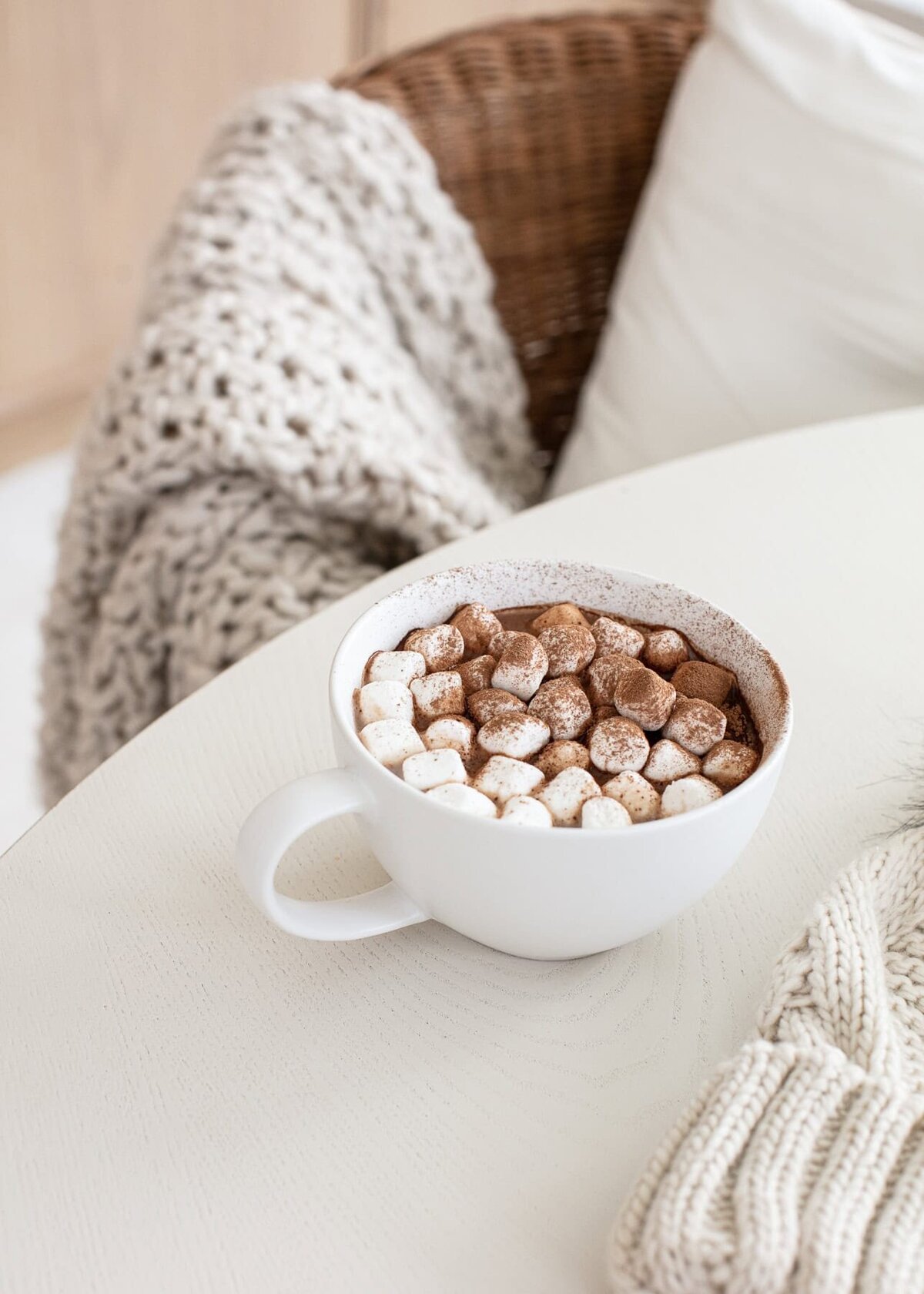 A cup of hot chocolate in a white mug with lots of marshmallows by a char with a sweater draped over it.