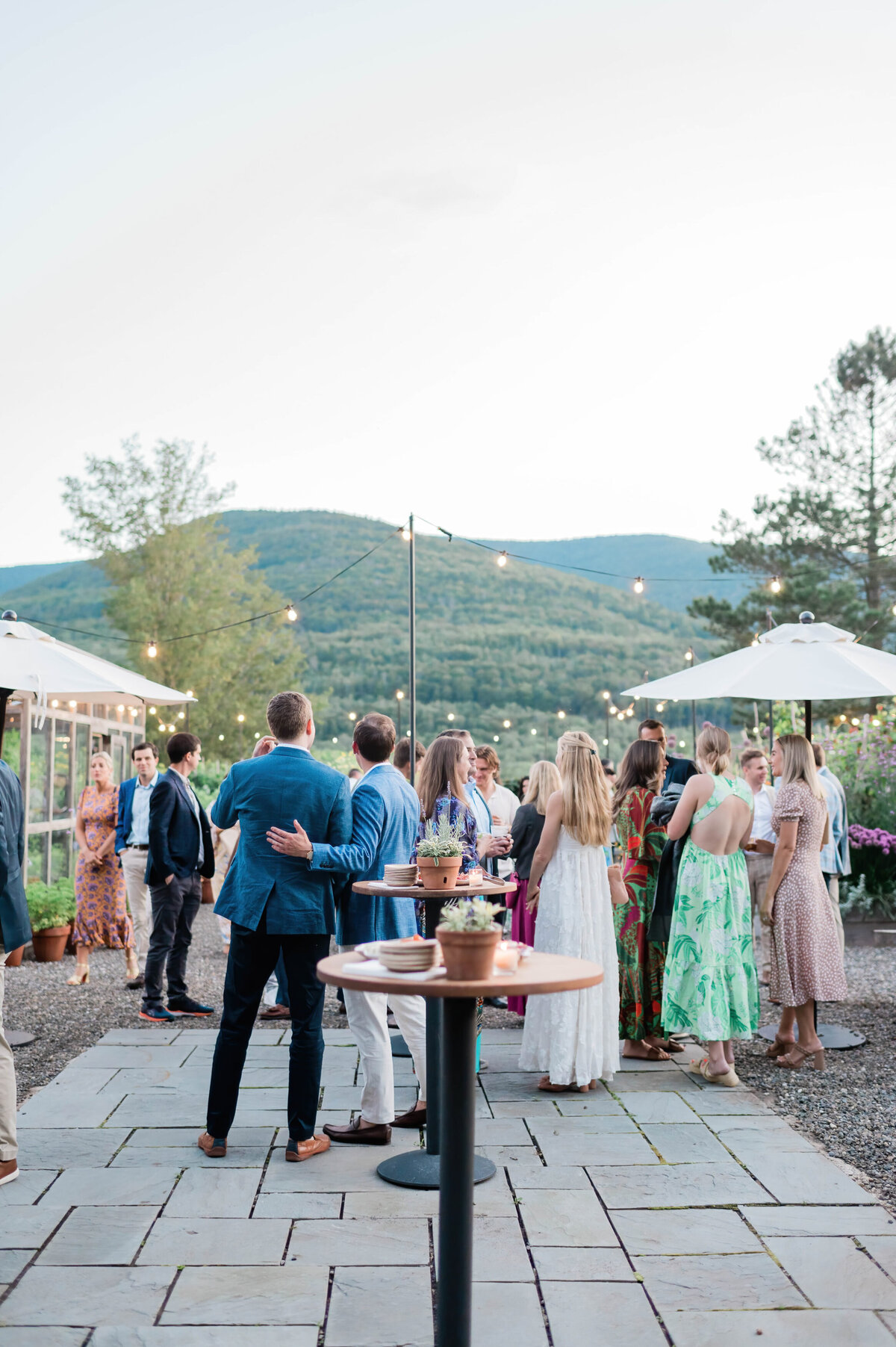  A group of guests, dressed in colorful attire, mingles under string lights with scenic mountain views at dusk, surrounded by tables and umbrellas.