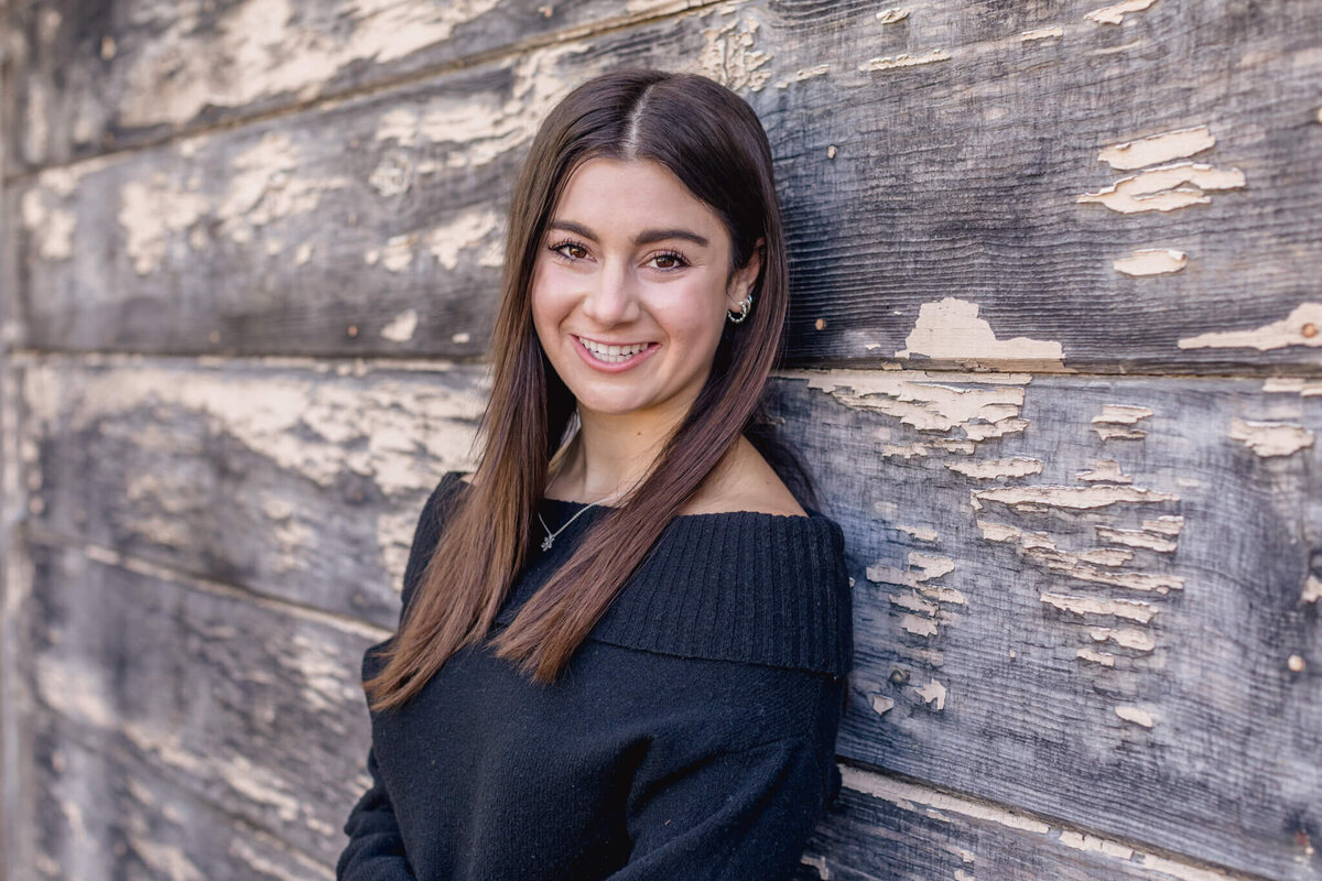 A close-up of a young woman with long, brown hair in a black sweater smiling sidelong at the camera and leaning against a warped wood wall.