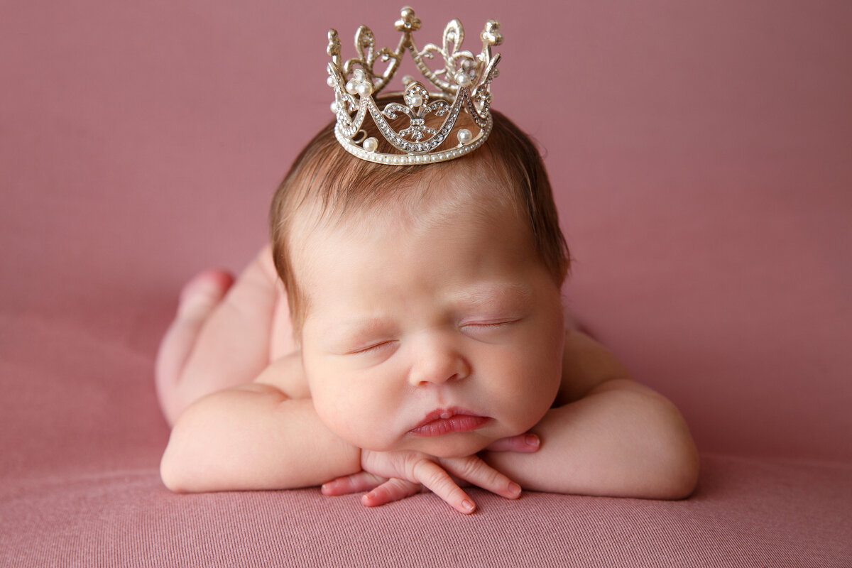 Portrait of a newborn baby girl sleeping on her tummy and lying on a prink blanet and wearing a tiny crown