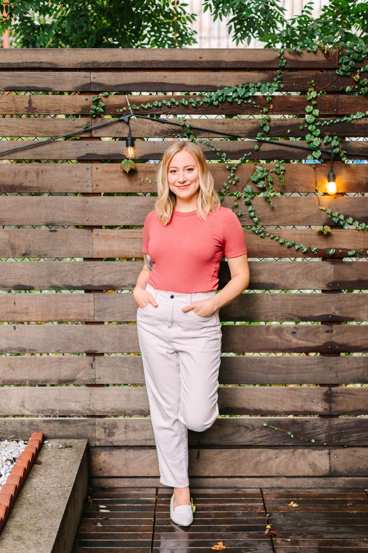 A woman with blonde hair and a warm smile leans against a wooden fence adorned with string lights and green vines. She wears a coral top, light-colored pants, and white flats, standing on a wooden deck.