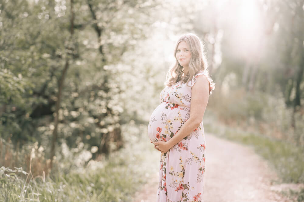 A beautiful pregnant mother smiles at the camera, as she is backlit by luminous glowing light during golden hour.