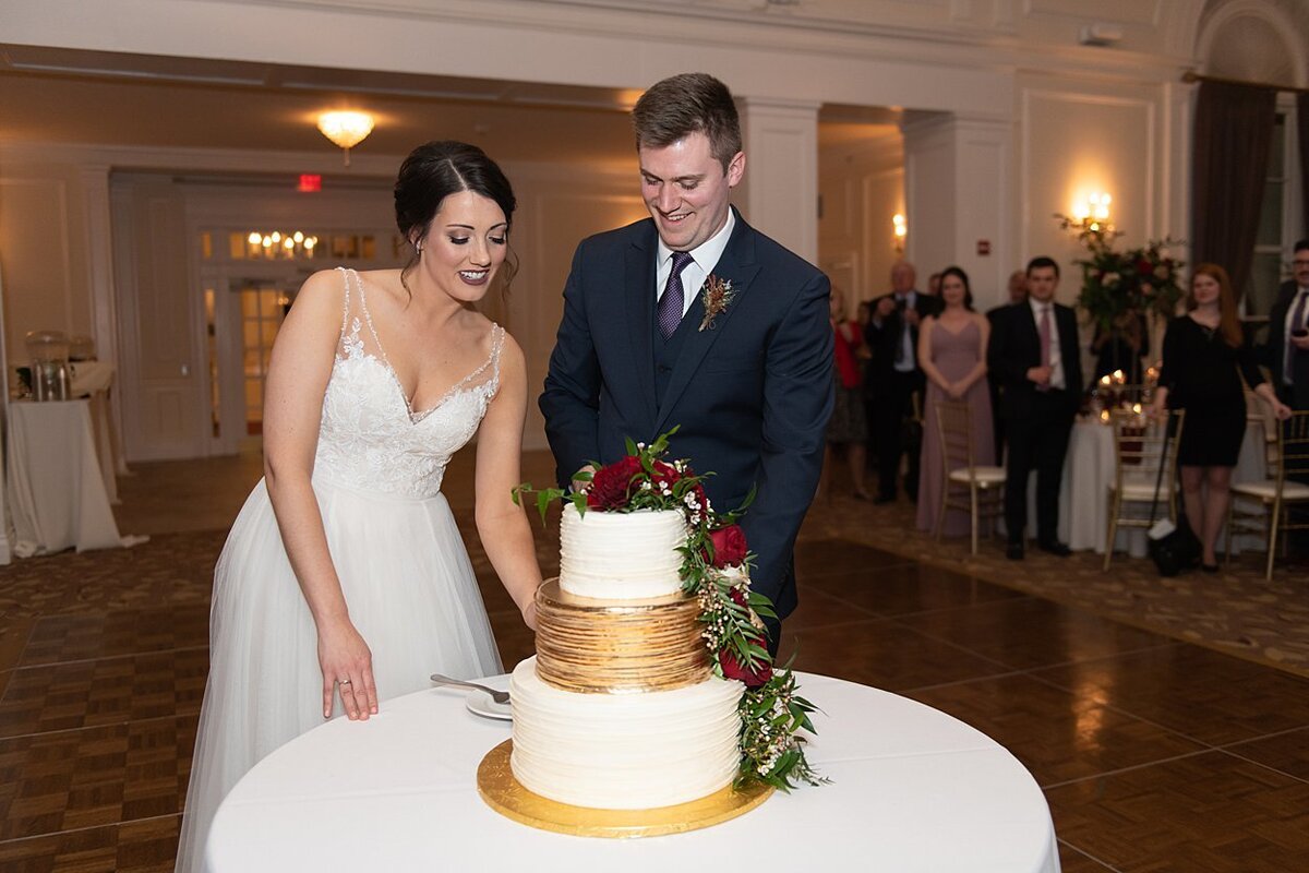 Bride and Groom cutting wedding cake with gold accents and red and off-white florals at University Club Ballroom in Pittsburgh, PA