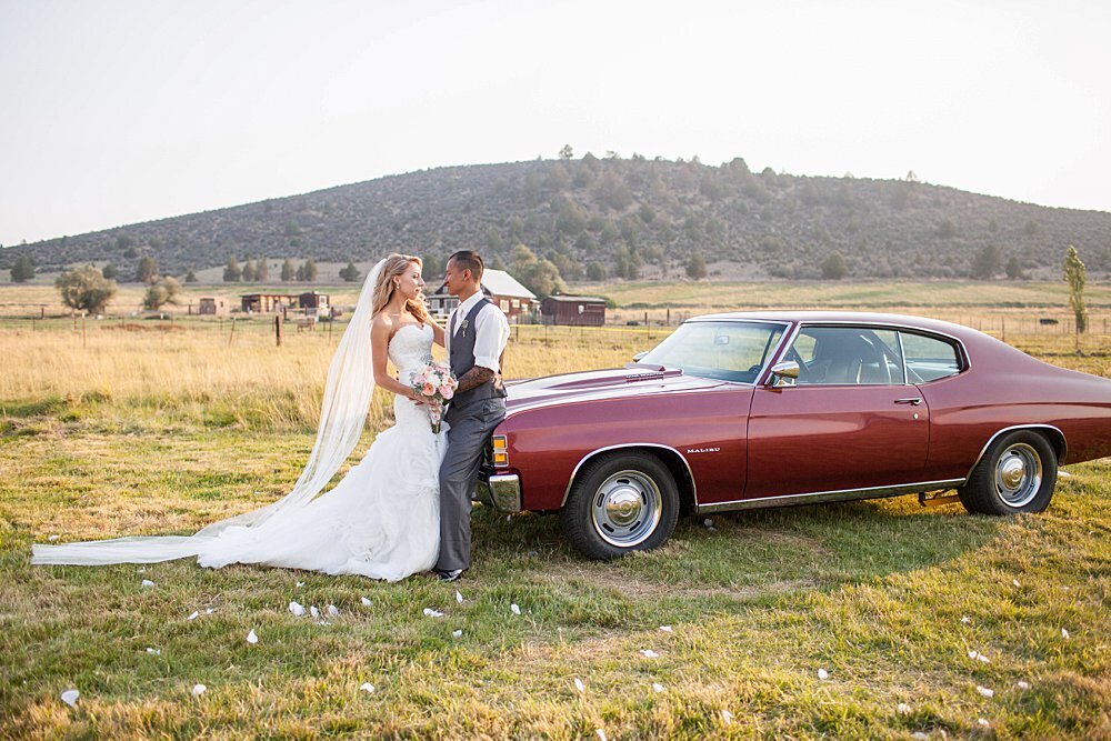 Newlyweds looking at each other next to maroon vintage car