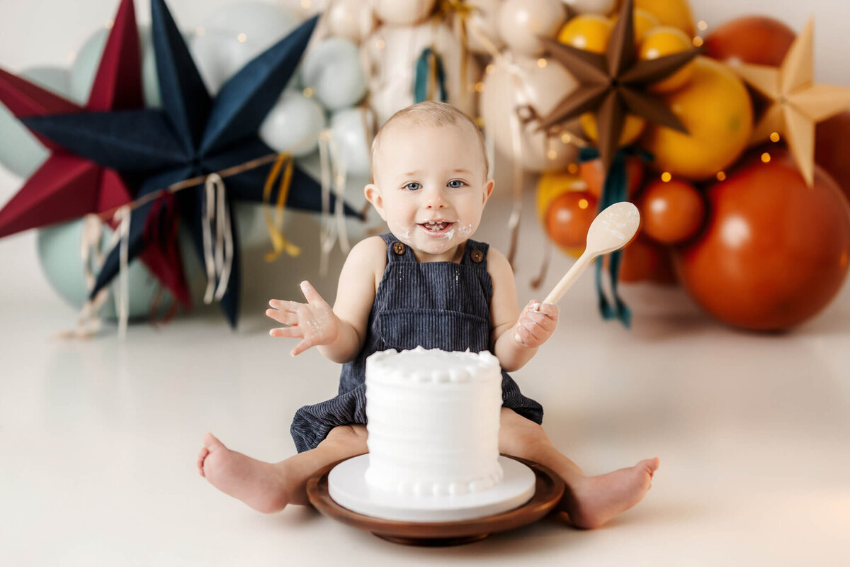 A baby in a dark romper smiles and holds a wooden spoon while sitting on the floor with a white cake. The background, crafted by a portrait photographer in Bloomington, Illinois, features colorful star-shaped decorations and balloons. Some cake is on the baby's face and hands.