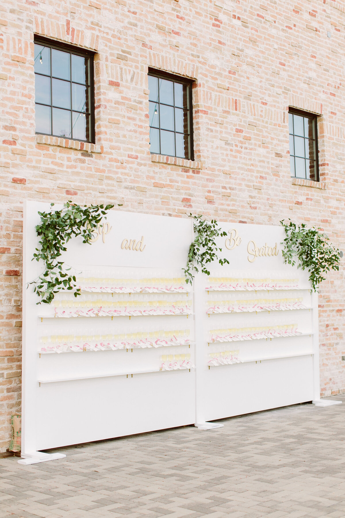 Large white wedding reception champagne backdrop display decorated with greenery against a brick wall