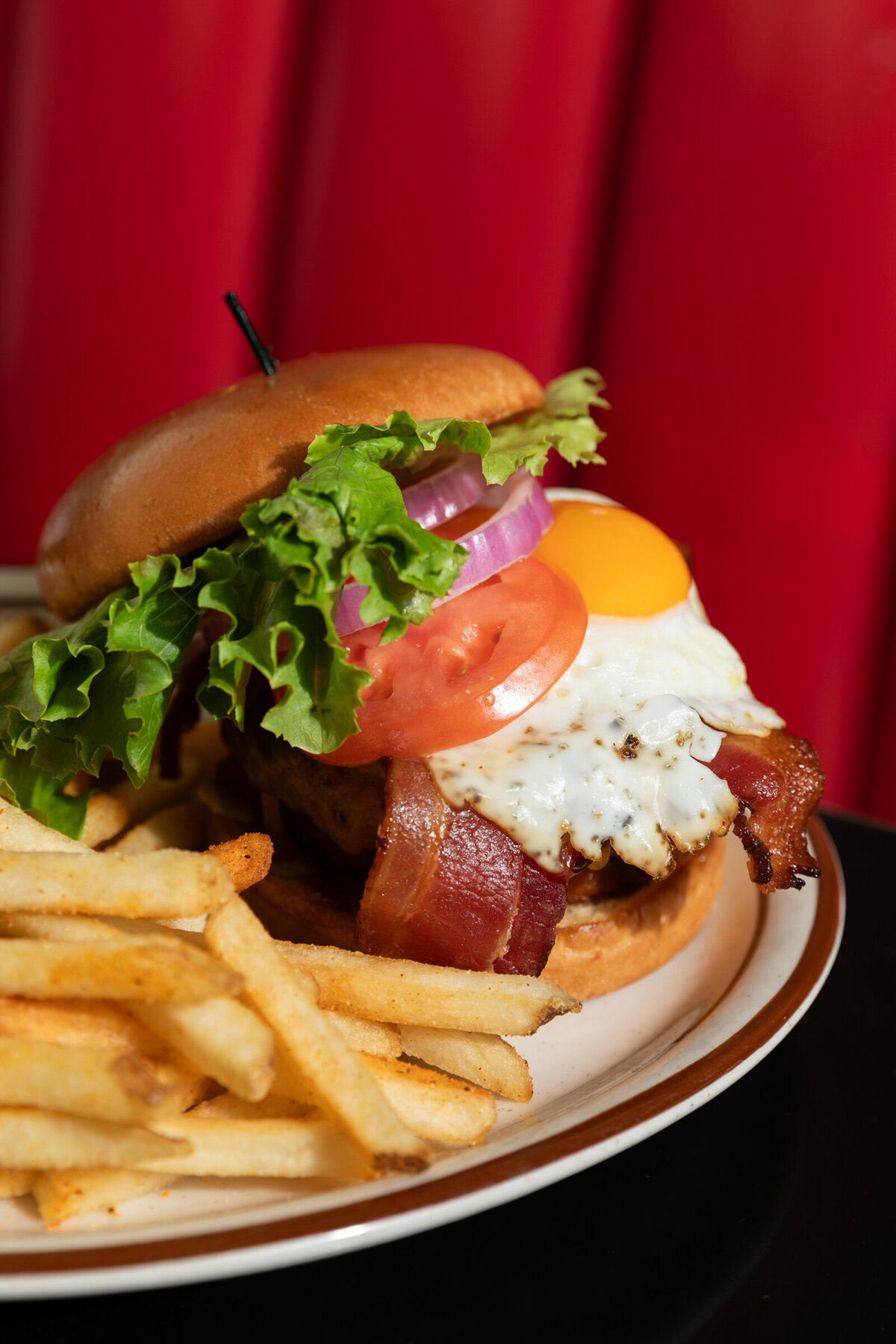 A close up image of a cheese burger with bacon, lettuce, tomato, onions, and a fried egg on a plate with fresh french fries.