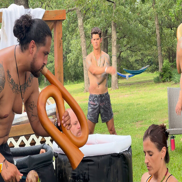 An image of a veteran retreat co-facilitator blowing into a horn while a woman and a man sitting inside an ice bath. A man prepares for the ice bath in the background.