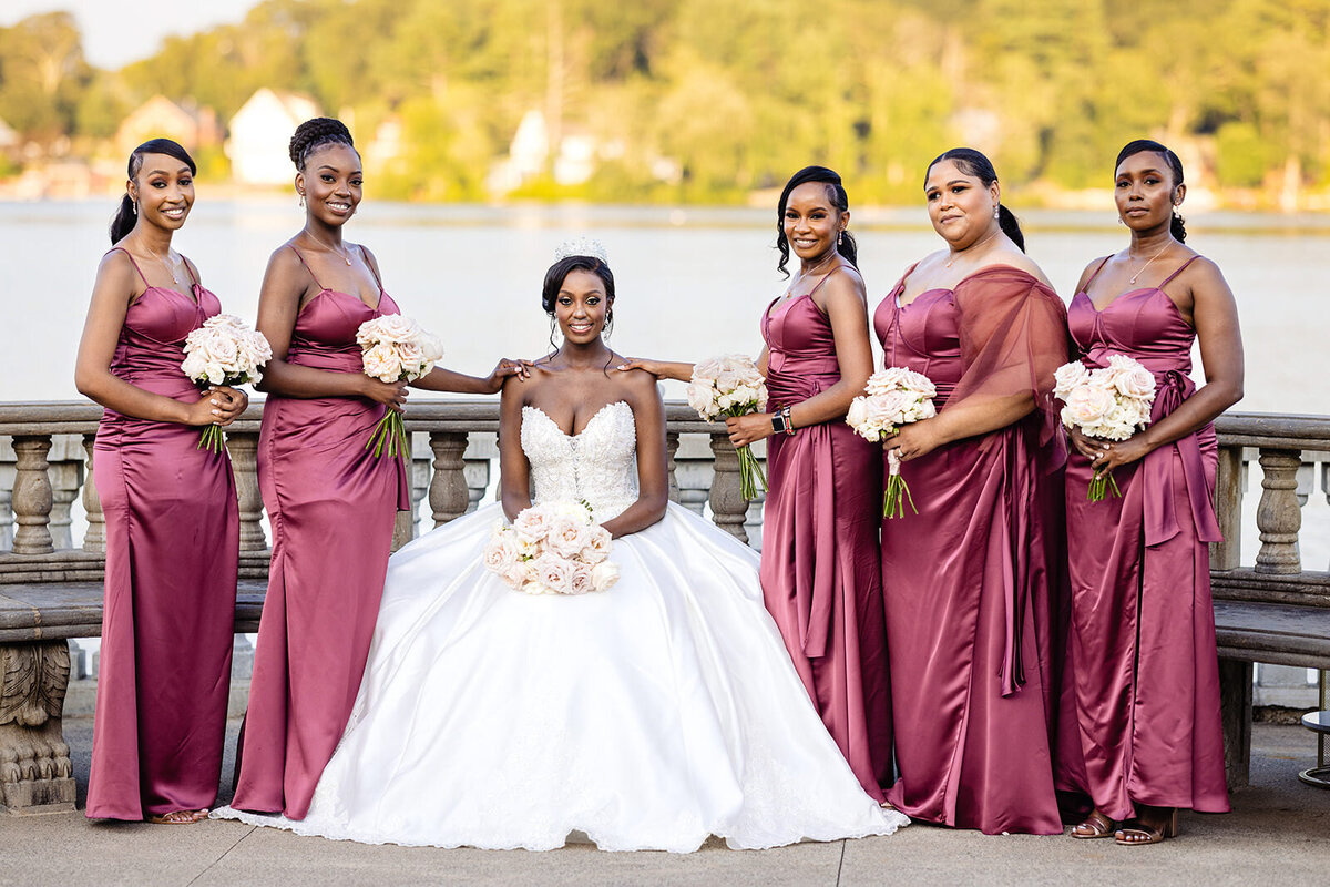 Bride seated and surrounded by bridesmaids standing in burgundy dresses, all holding bouquets by a lakefront