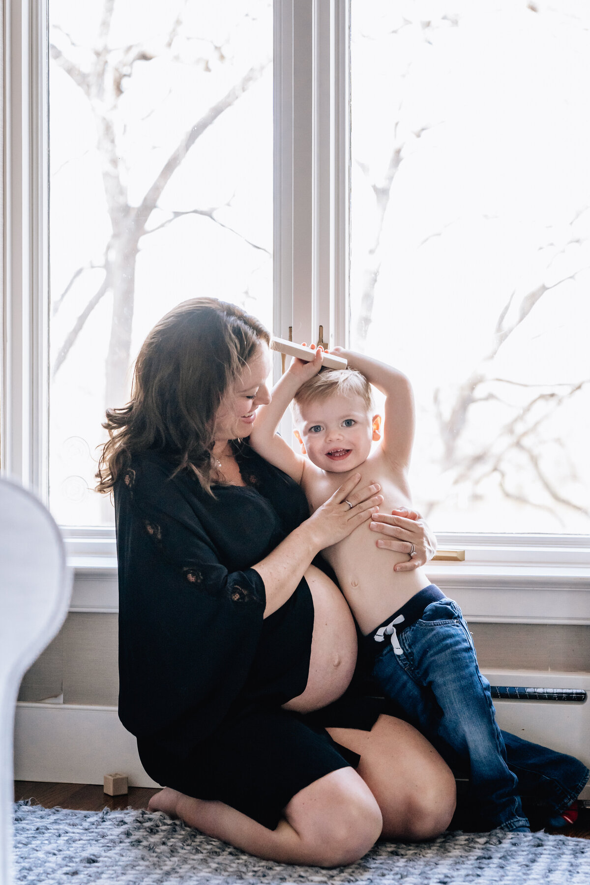 A pregnant woman wearing a black dress sits on the floor by a large window, lovingly holding and smiling at her young son who is playfully stretching his arms above his head.