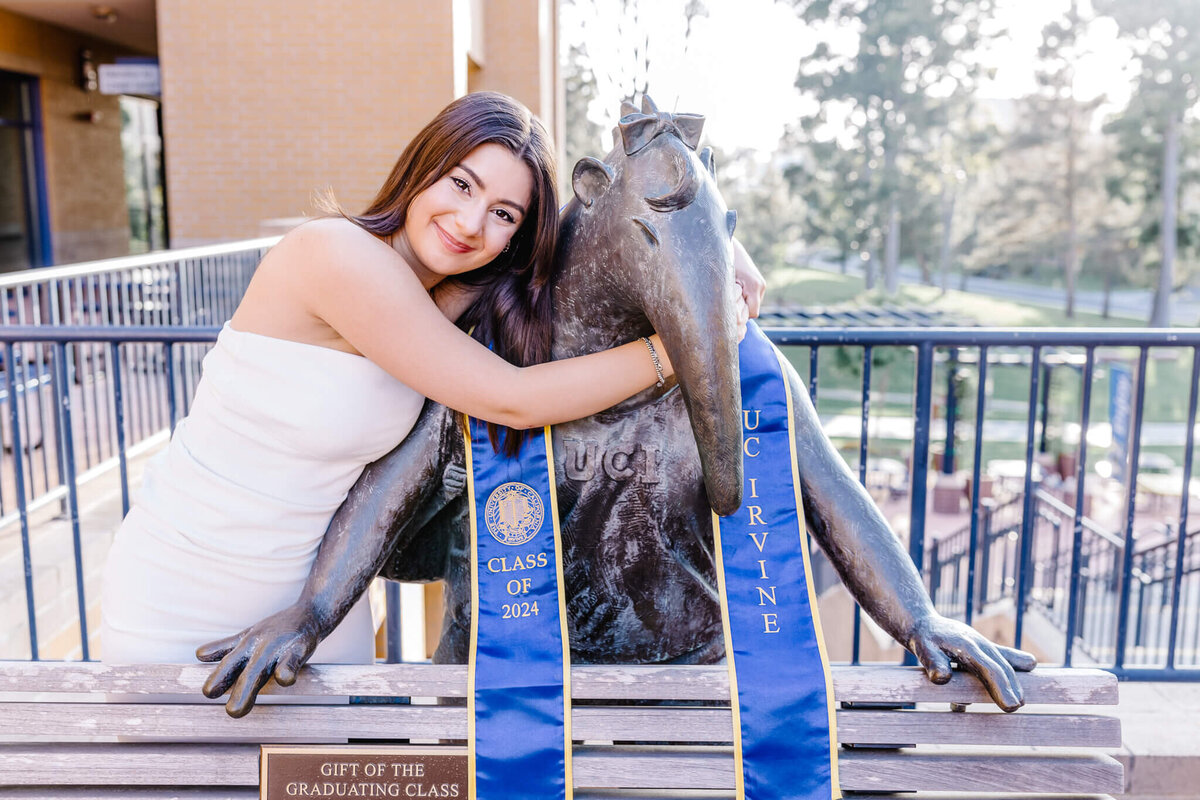 A young, female college graduate with brown hair wearing a white dress hugging a bronze statue of an anteater, the UCI mascot, with a dark blue graduation sash around the statues neck. Captured at the University of California, Irvine.