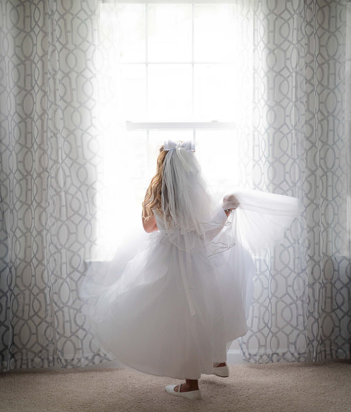 Little girl is twirling in her first communion gown in front of a window. Her back is to the camera. Captured by Liza Nicole Photography, Philadelphia and Main Line family photographer.