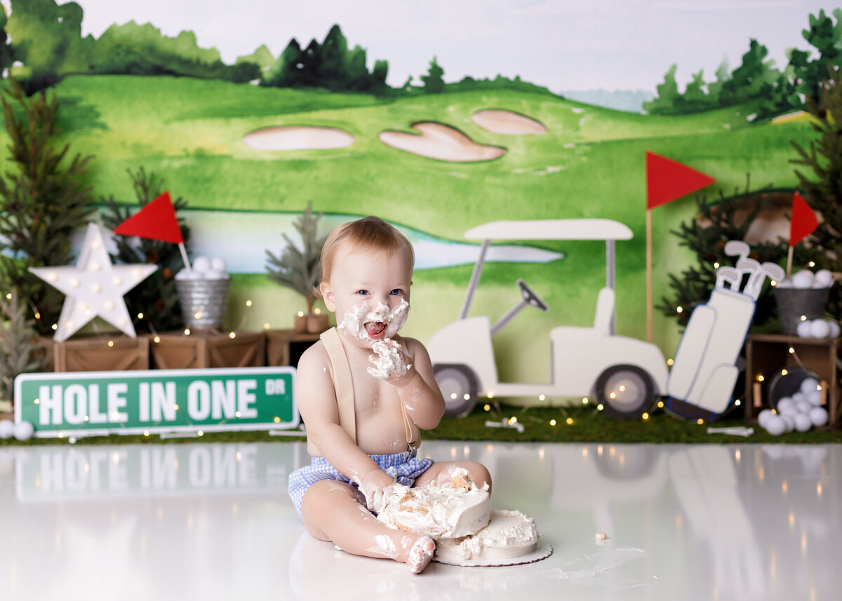 Rustic golf themed cake smash in Palm Beach and Lake Worth Florida photography studio. Baby boy is wearing light blue diaper cover with tan suspenders and has frosting on his hands, face and feet.  In the background is a golf theme backdrop with trees, golf cart and various golf decorations with scattered golf balls.