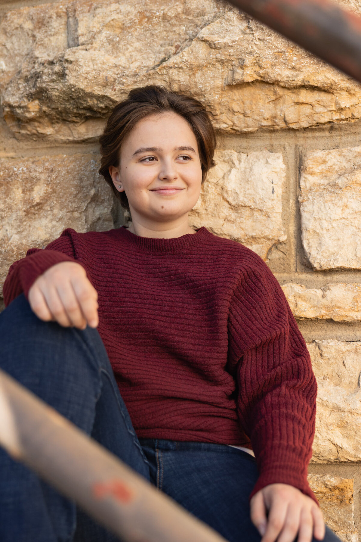 Senior girl sitting against a stone wall wearing a red sweater during fall senior photos in Lawrence KS