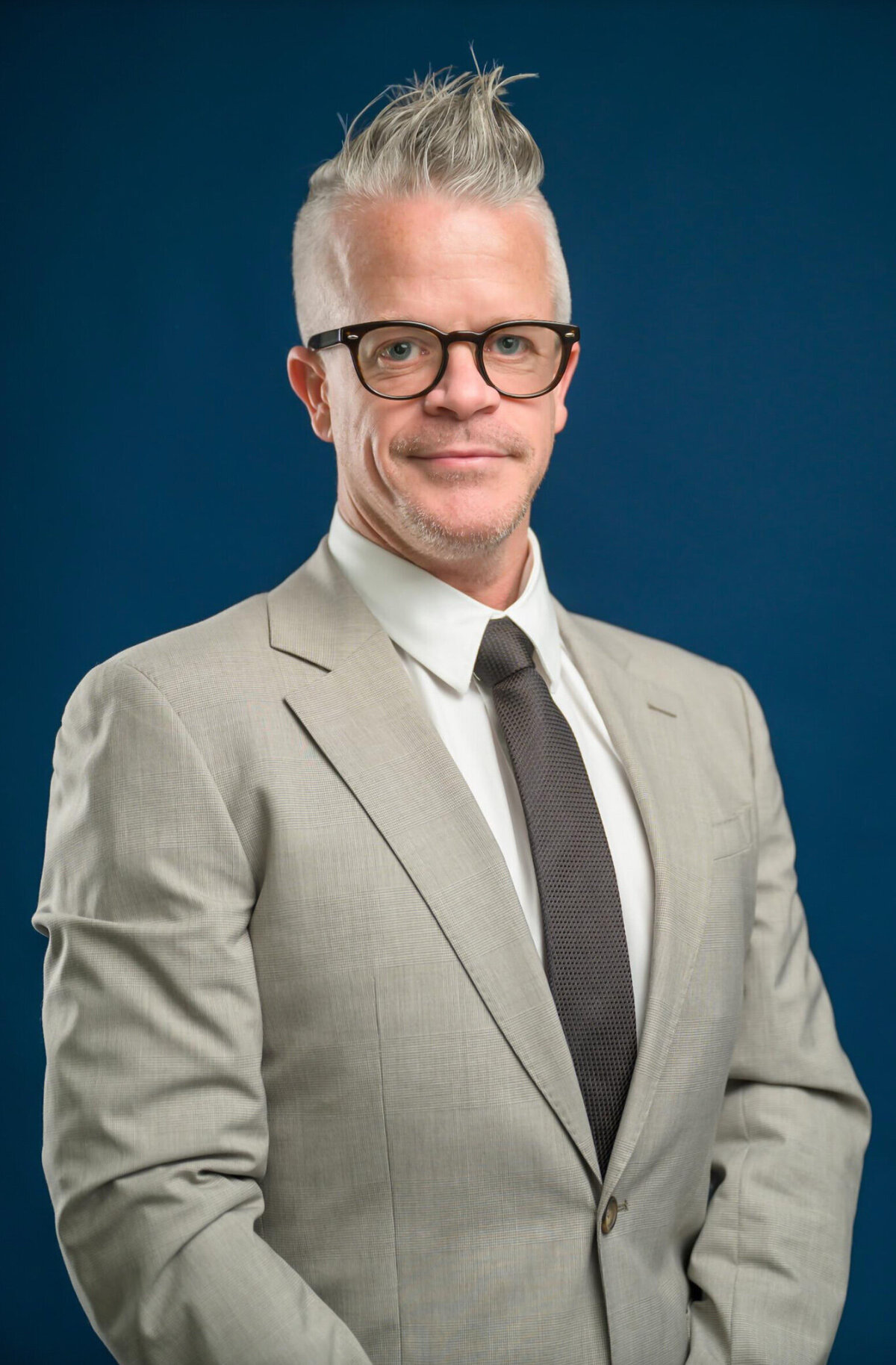 Man with spiky gray hair, wearing a light gray suit posing for a headshot in front of a blue background