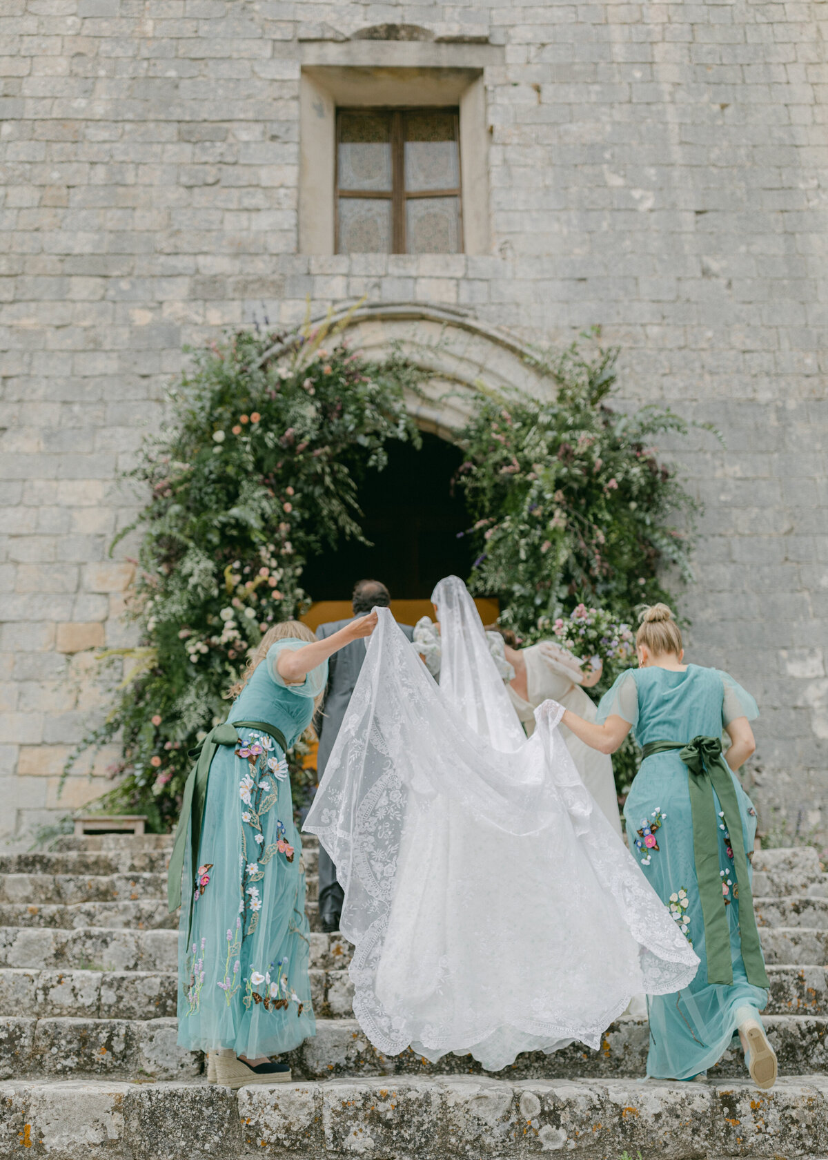 Bride walking up church stairs with bridesmaids helping her