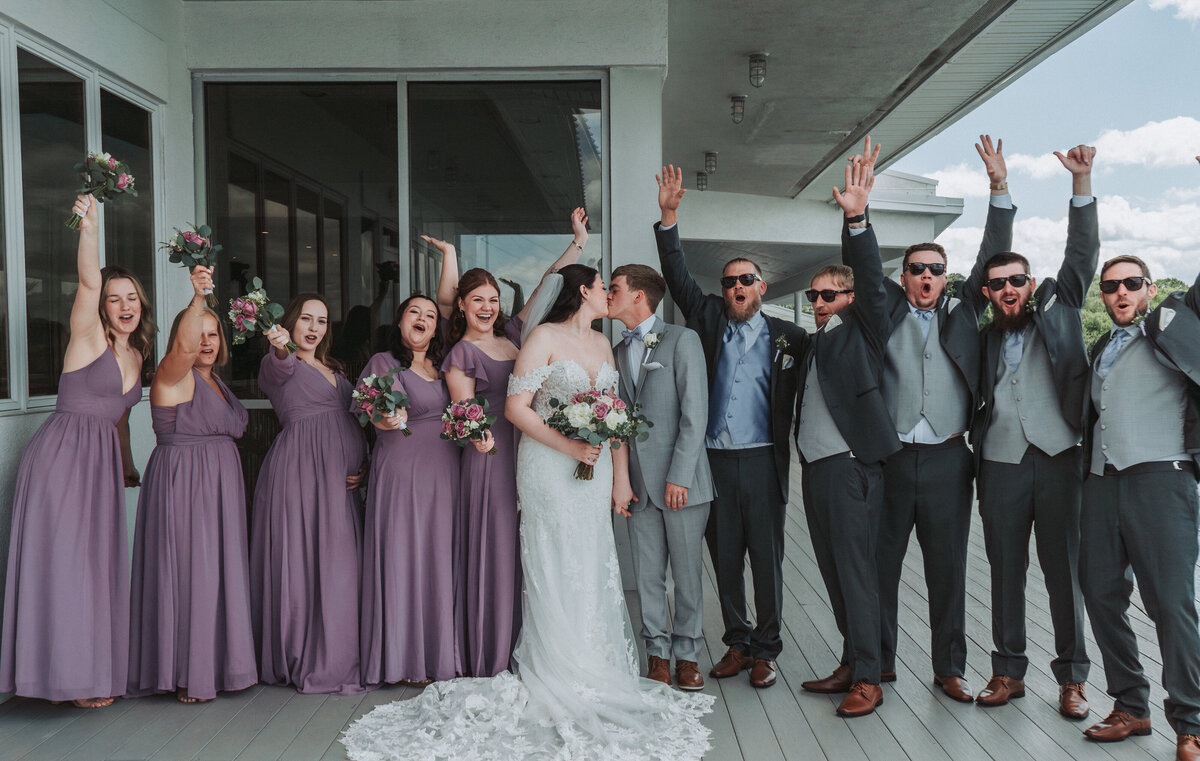 Bride and groom kiss while their bridal party in lavender and gray cheers with arms raised outside the Lesner Inn wedding venue, Virginia Beach Virginia.