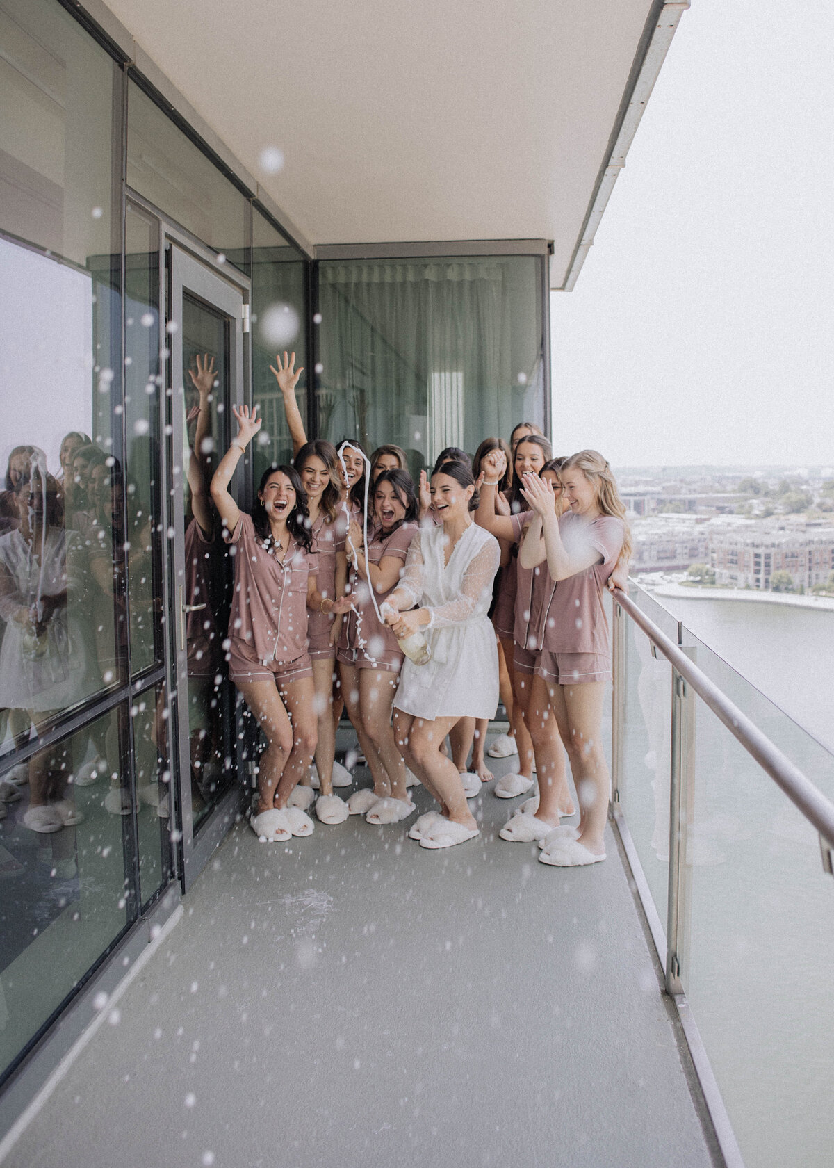 Bride and her bridesmaids celebrate by popping champagne on a high-rise balcony, laughing as bubbles spray around them.
