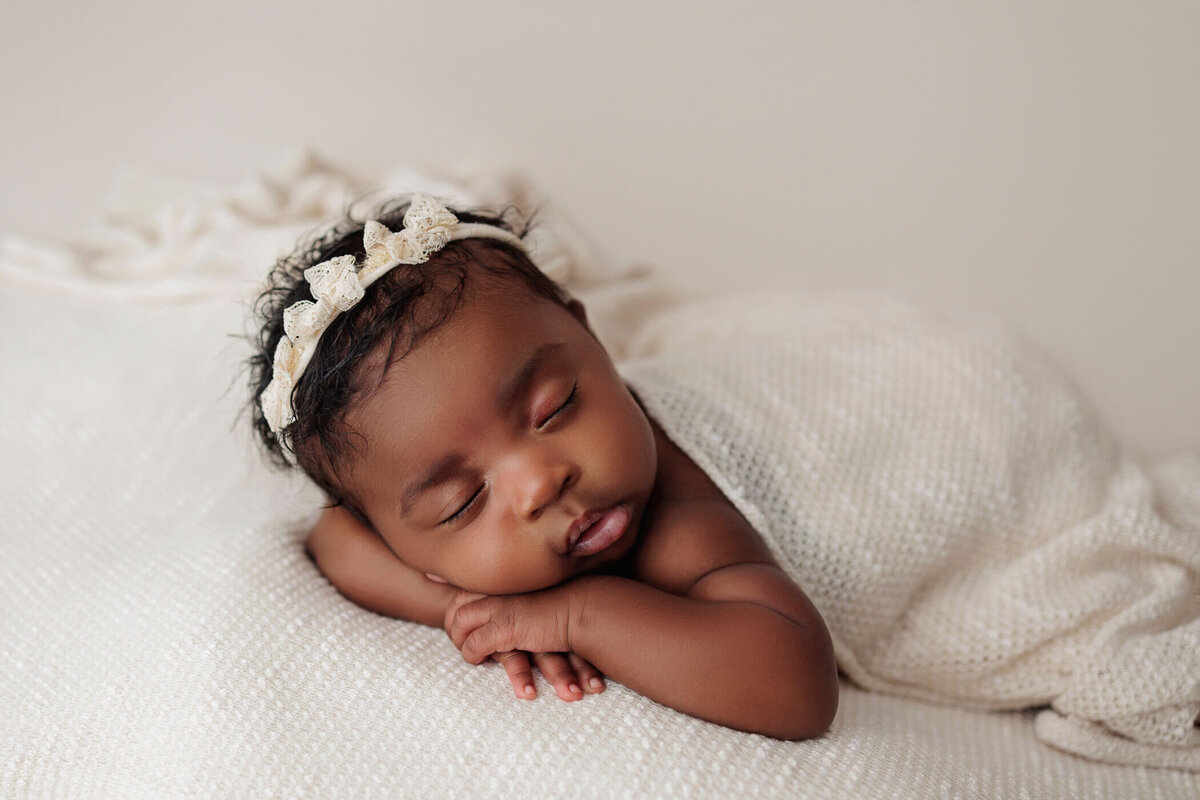 Newborn baby girl sleeping peacefully on her stomach, wrapped in a soft white blanket, with a white headband adorned with a bow.