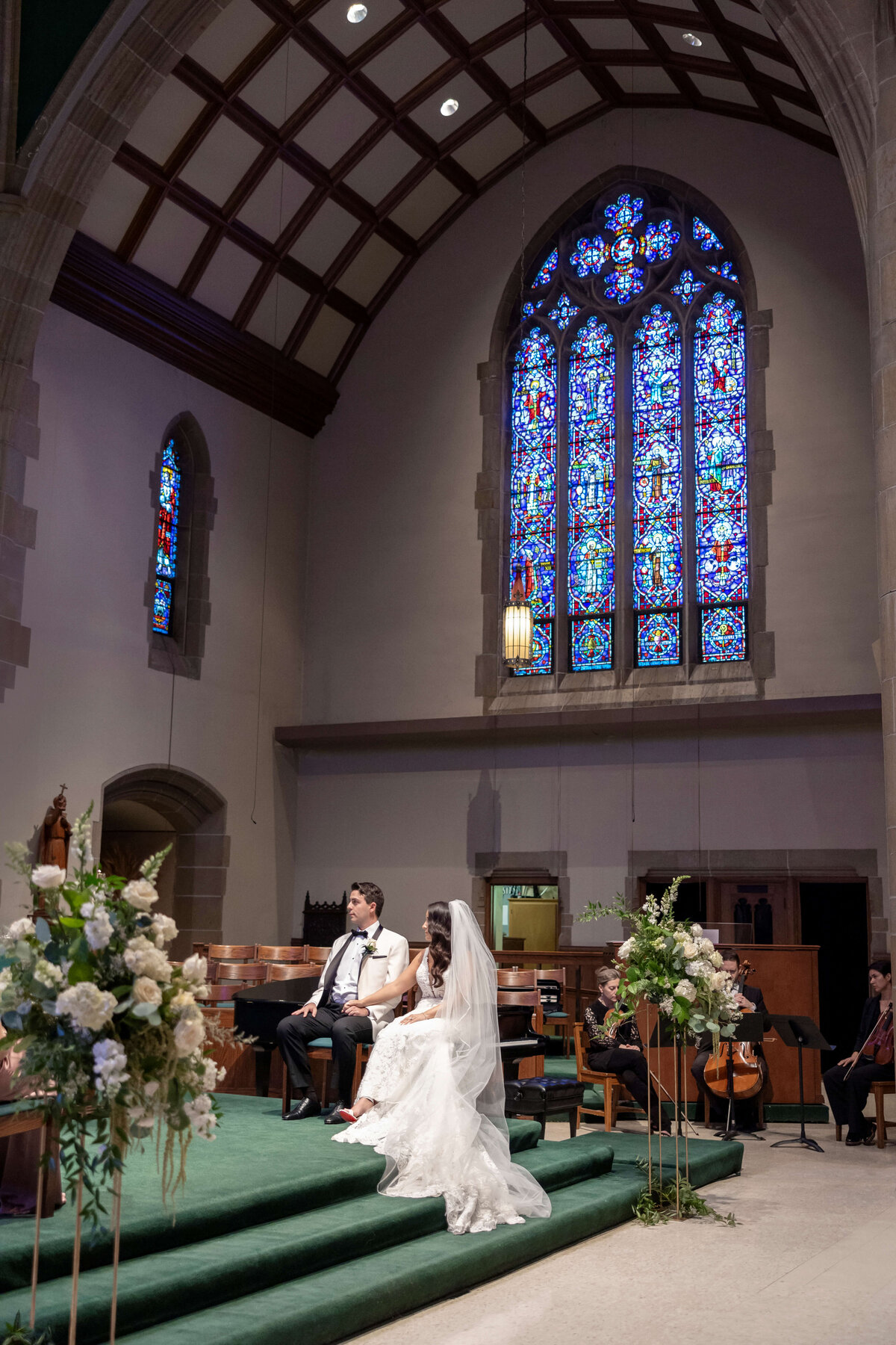 A bride and groom sit on a stage in a church, beneath a large stained glass window. The bride wears a white gown and veil, while the groom is in a dark suit. Floral arrangements decorate the stage, and musicians play in the background.