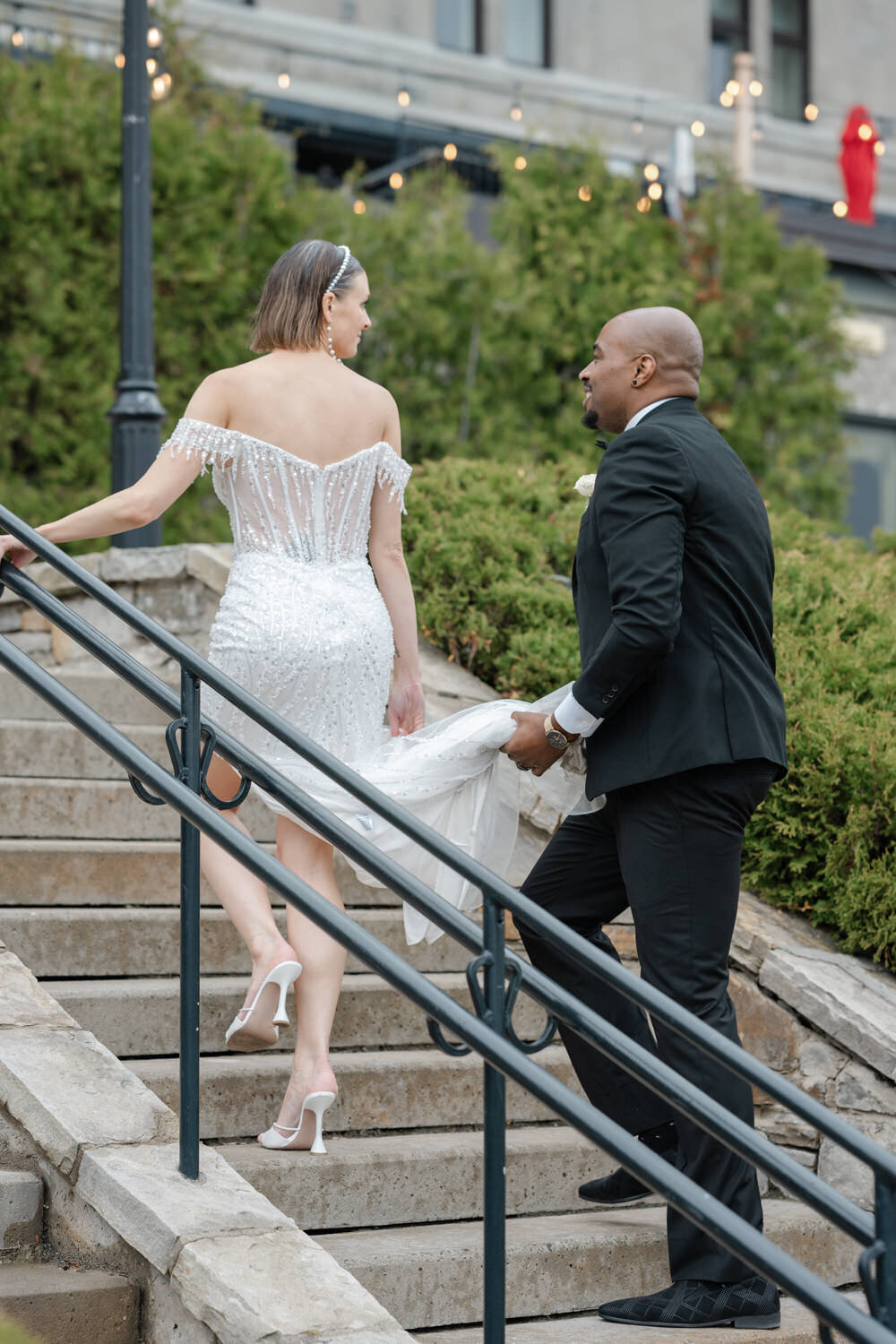 Le marié assiste tendrement sa femme en tenant sa robe lors d’un mariage de prestige à Montréal, geste complice capturé dans une ambiance chic et attentionnée