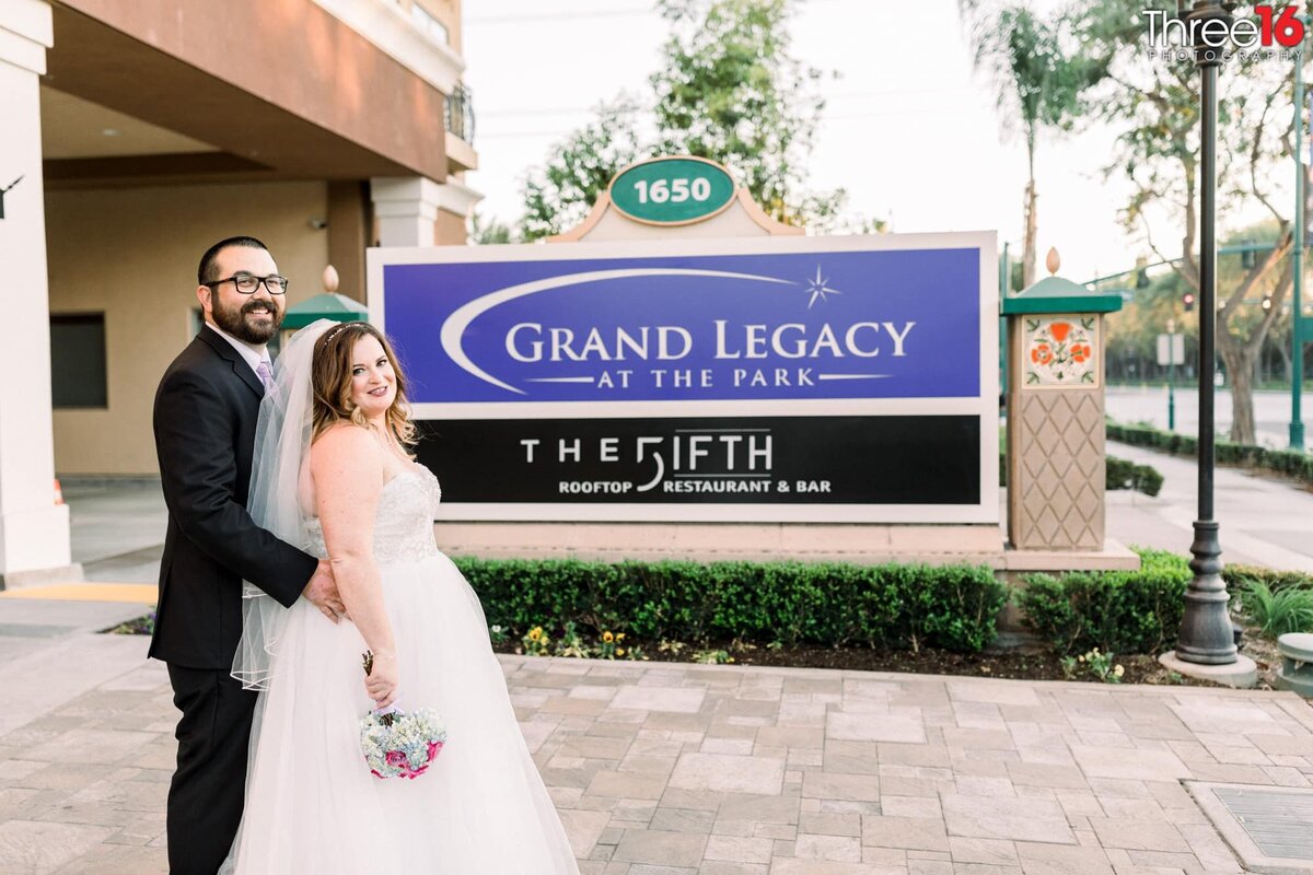 Groom holds his Bride from behind as they pose in front of the Grand Legacy signage