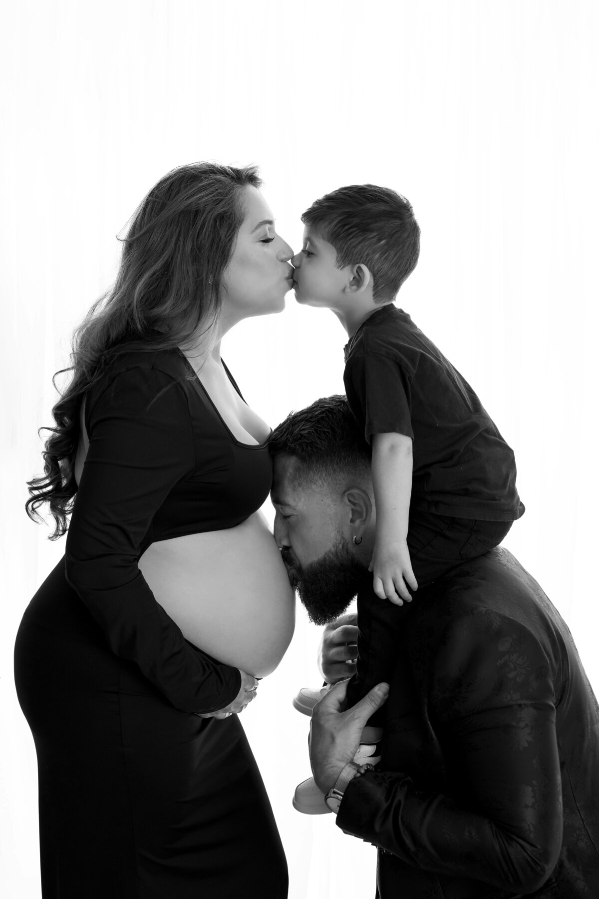 Expecting mom kisses son while dad carries him on shoulders and kisses her pregnant belly.