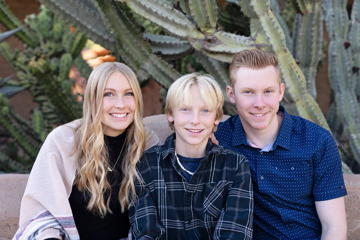 Three blonde siblings (two brothers and their sister) sitting together and smiling at the camera with cactus plants behind them.